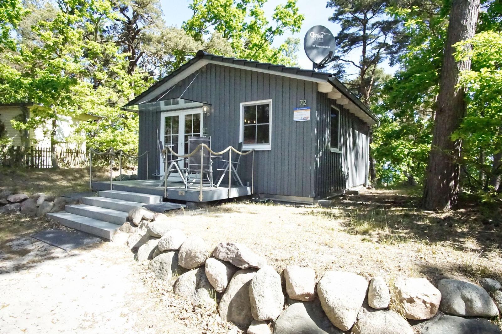 Graues Chalet mit Terrasse, Satellitenantenne und Umgebung aus Bäumen und Steinen.