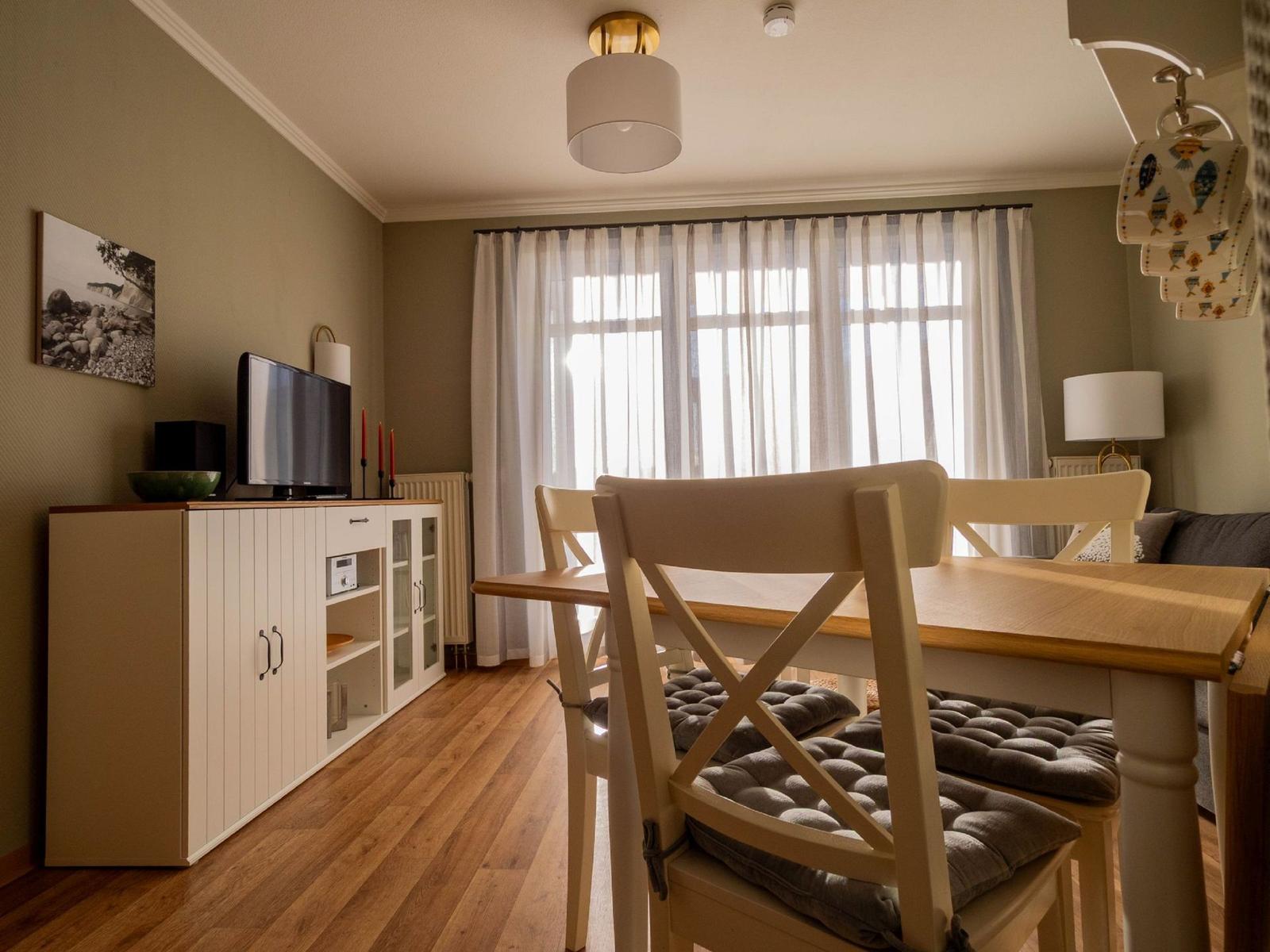 Esszimmer mit Holztisch, Stühlen und TV-Schrank. Fenster mit Vorhängen.