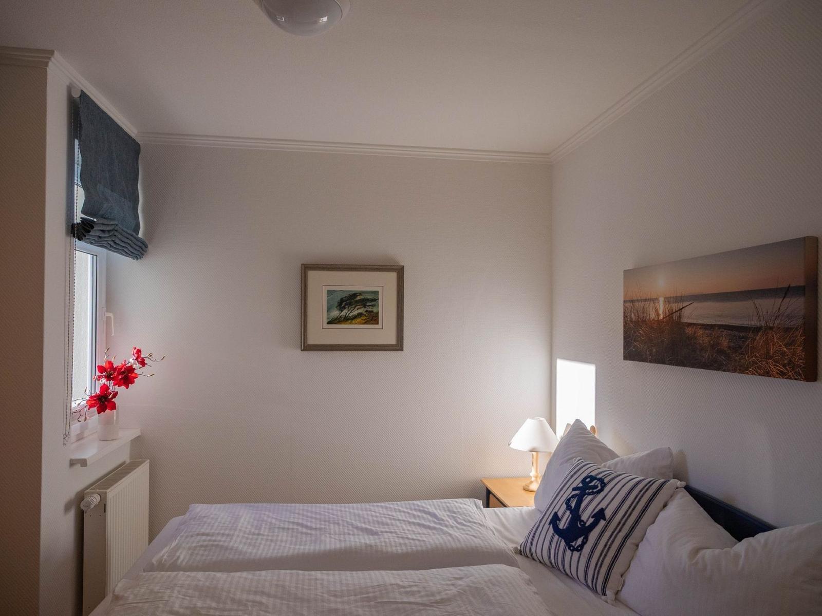 Schlafzimmer mit Bett, Nachttisch, Wandbild und Fenster mit Blumen.
