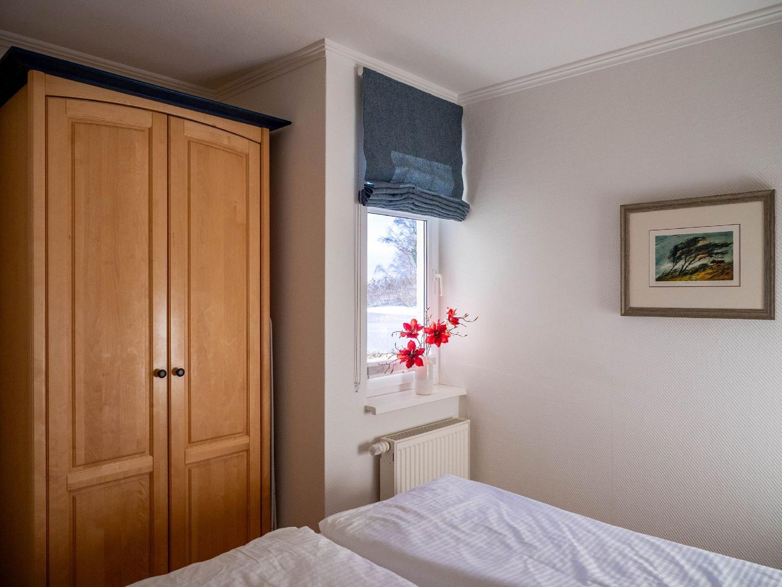 Schlafzimmer mit Holzschrank, Fenster mit blauem Rollladen und Blumen auf dem Fensterbrett.