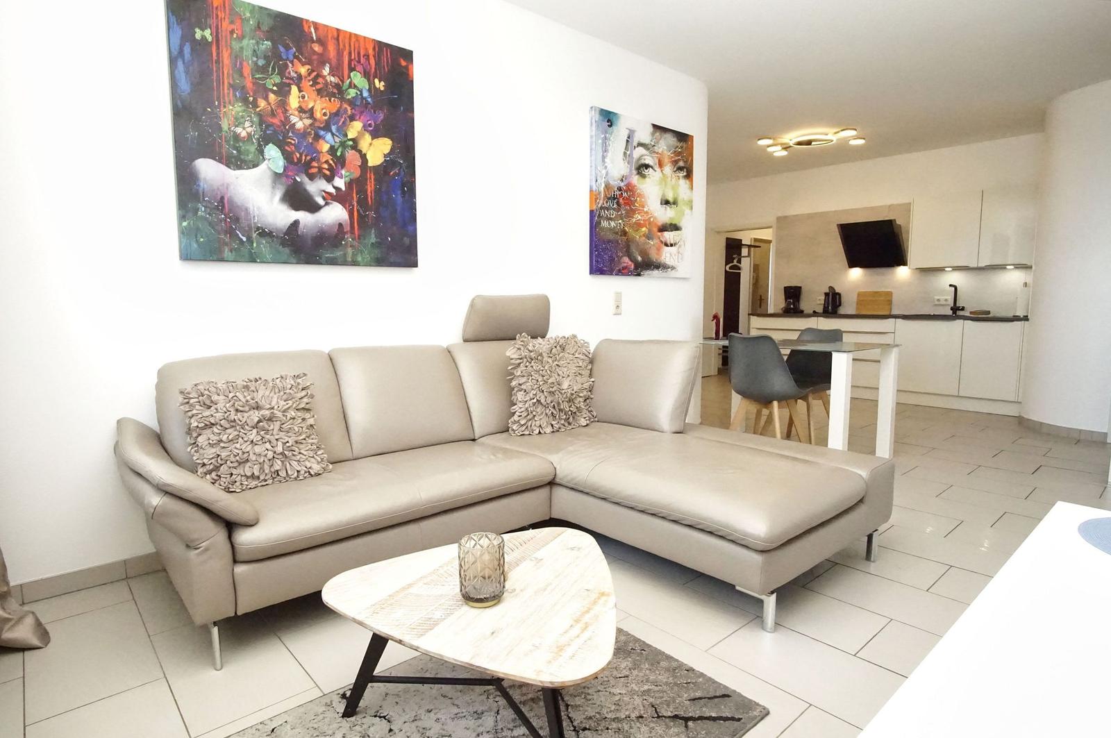 Wohnbereich mit Ecksofa, Couchtisch und Wandbildern. Küche im Hintergrund.
