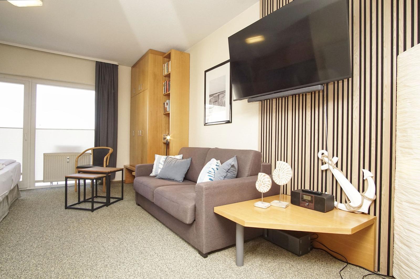 Wohnzimmer mit Sofa, Fernseher und Tisch. Fenster und Bett im Hintergrund.