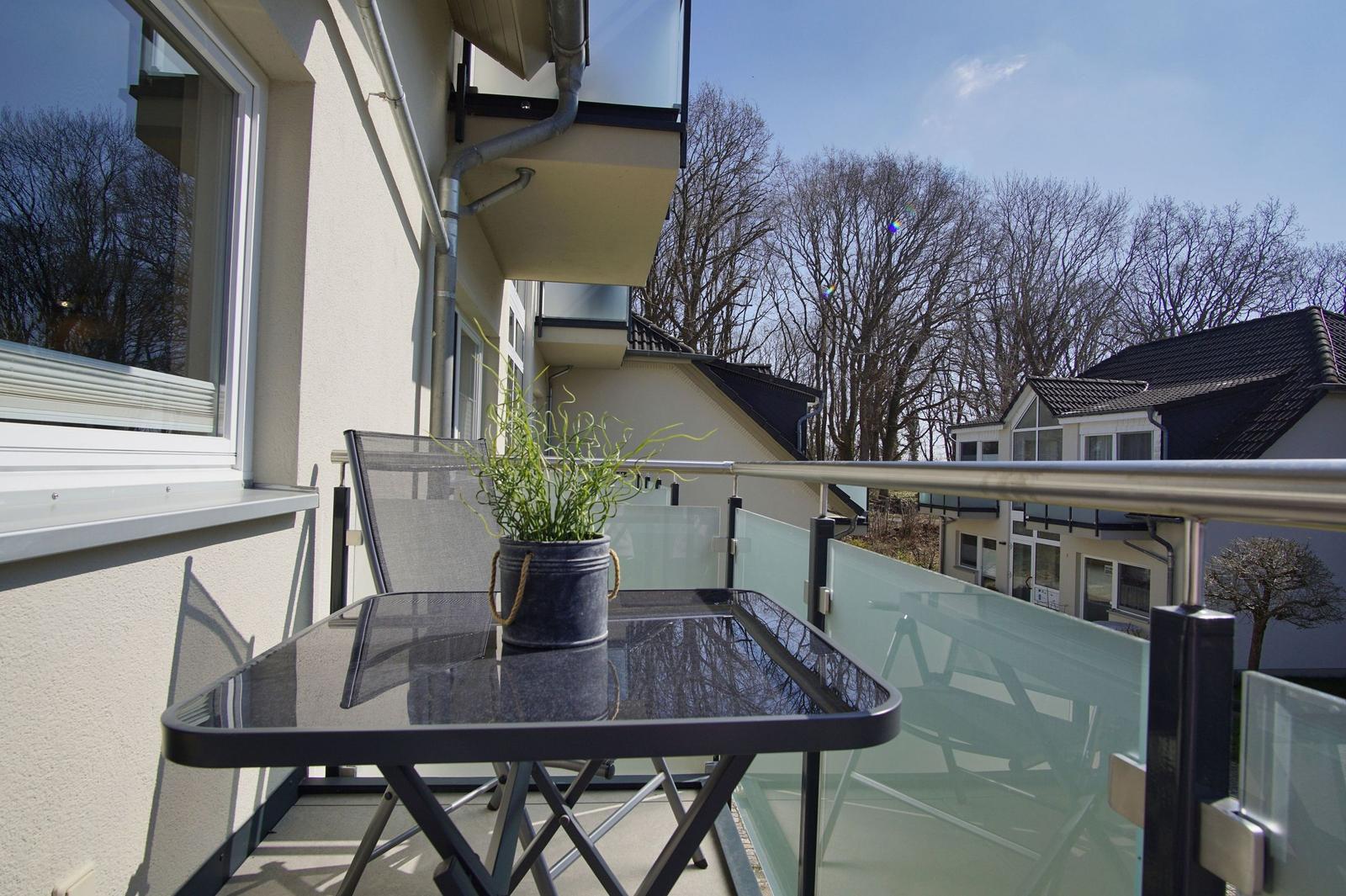 Terrasse mit Tisch, Stuhl und Pflanze. Glasgeländer und Blick auf Häuser und Bäume.