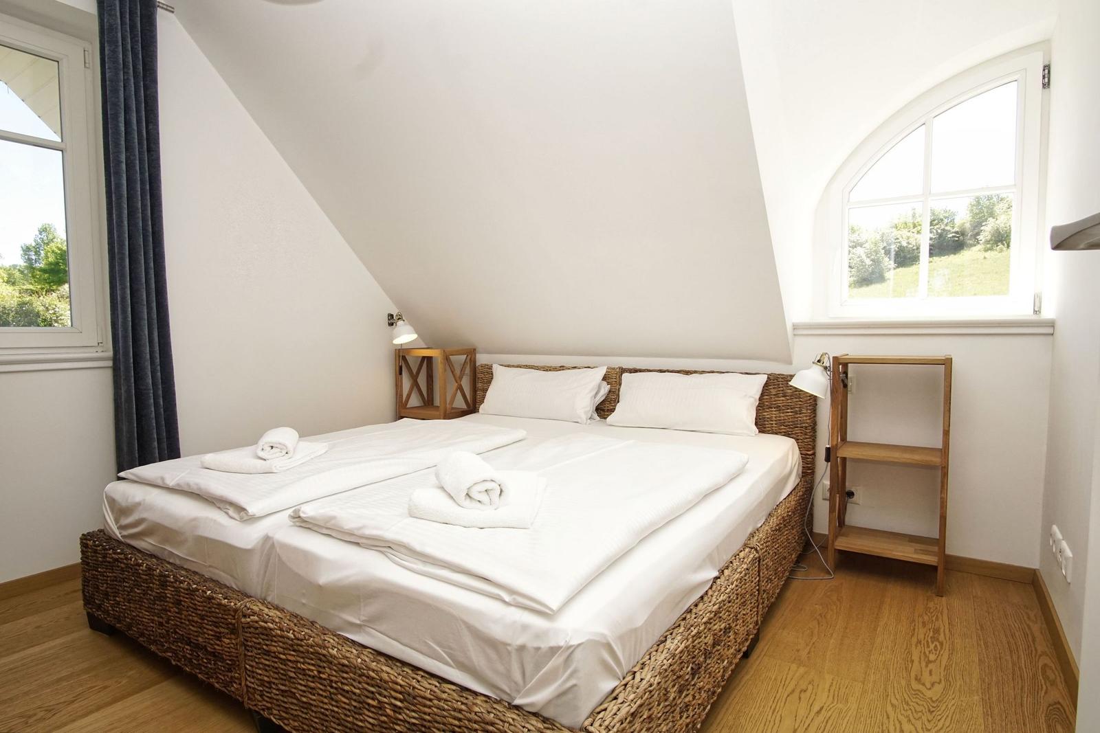 Schlafzimmer mit Doppelbett, Holzboden und zwei Fenstern.