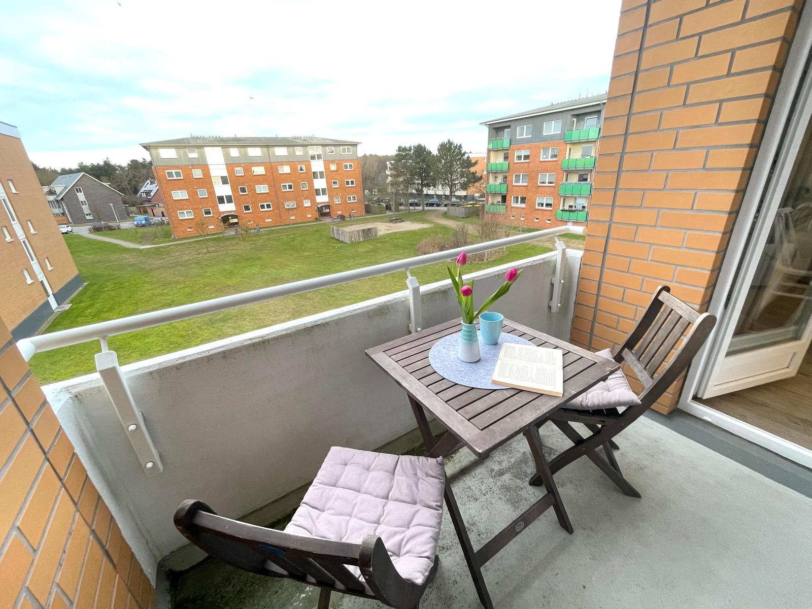 Balkon mit Tisch, zwei Stühlen und Blumen. Blick auf Grünfläche und Wohnhäuser.