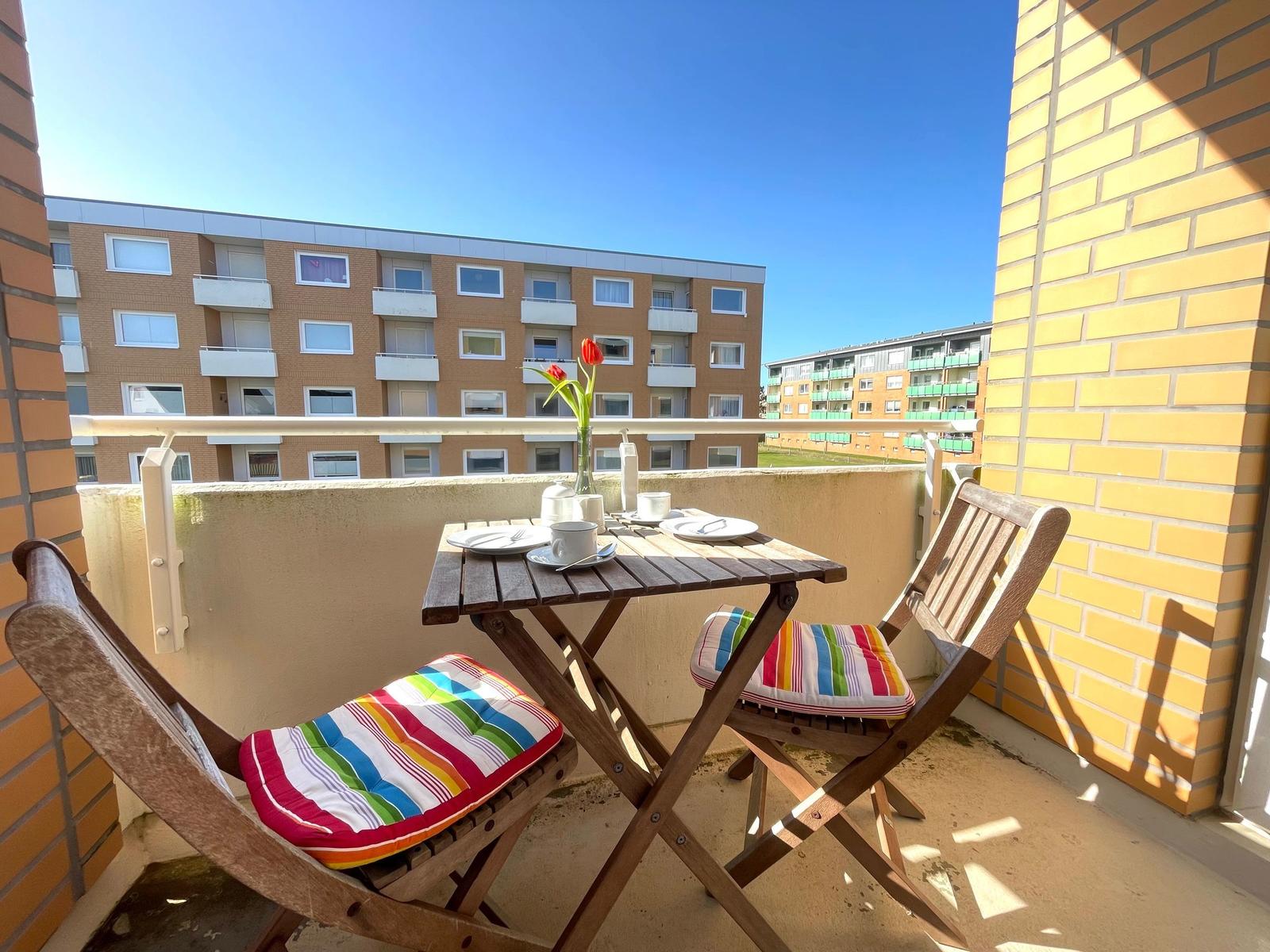 Balkon mit Tisch, zwei Stühlen und roter Tulpe. Hintergrund: Wohnhaus und klarer Himmel.