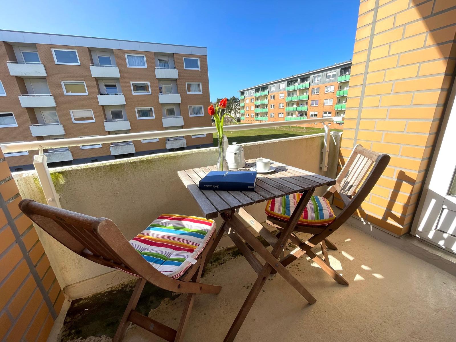 Terrasse mit Tisch, zwei Stühlen und Blumen. Blick auf Wohnhäuser und Grünfläche.