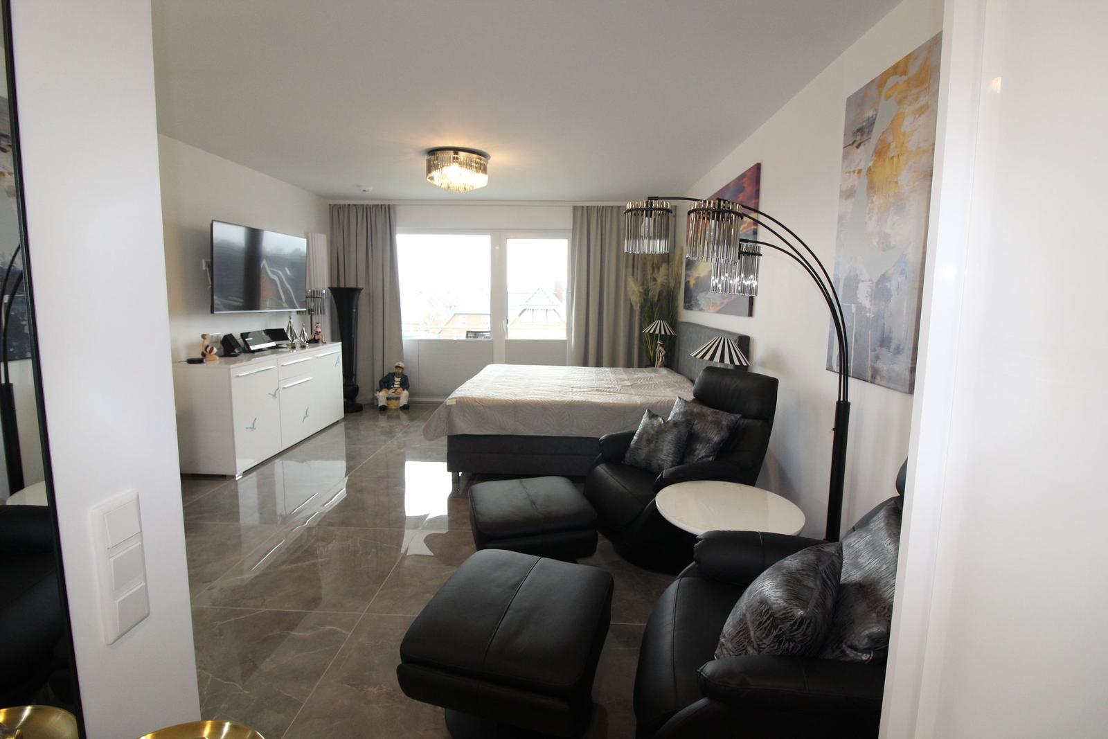Schlafzimmer mit Bett, Sessel und Fernseher. Große Fenster und moderne Beleuchtung.