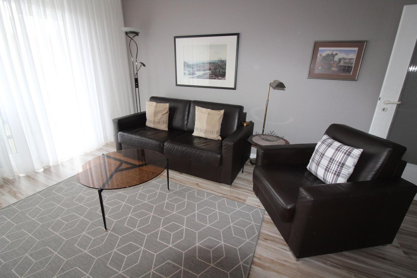 Wohnzimmer mit Sofa, Sessel, Glas-Tisch und Wandbildern.