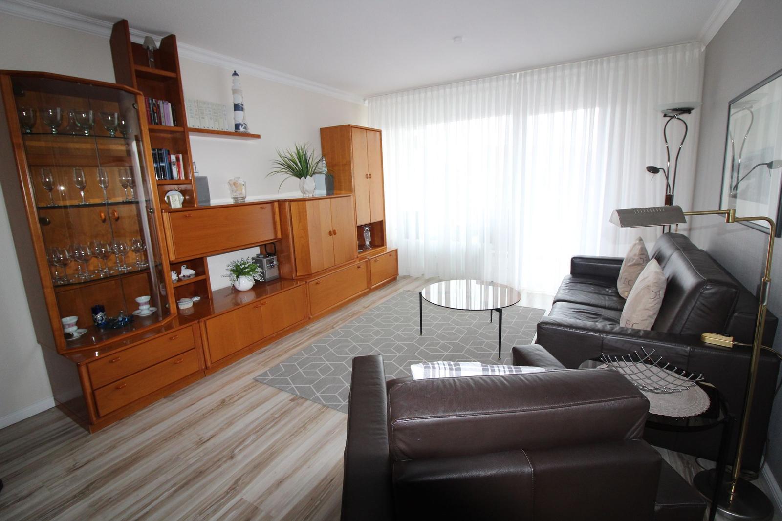 Wohnzimmer mit braunem Ledersofa, Holzmöbeln und Glasvitrine. Fenster mit weißen Vorhängen.