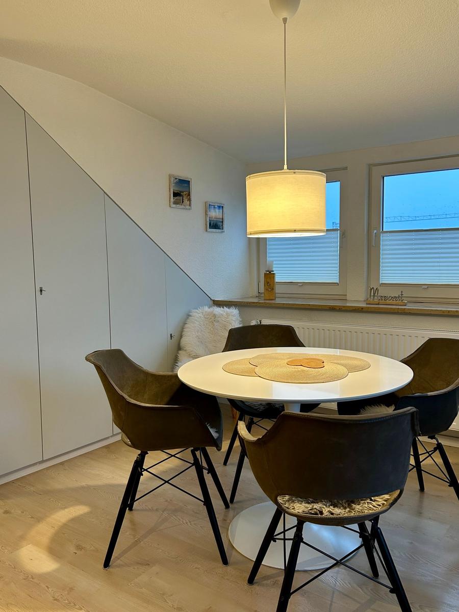 Runder Esstisch mit vier Stühlen unter einer Pendelleuchte. Fenster mit Jalousien und Wandregal.