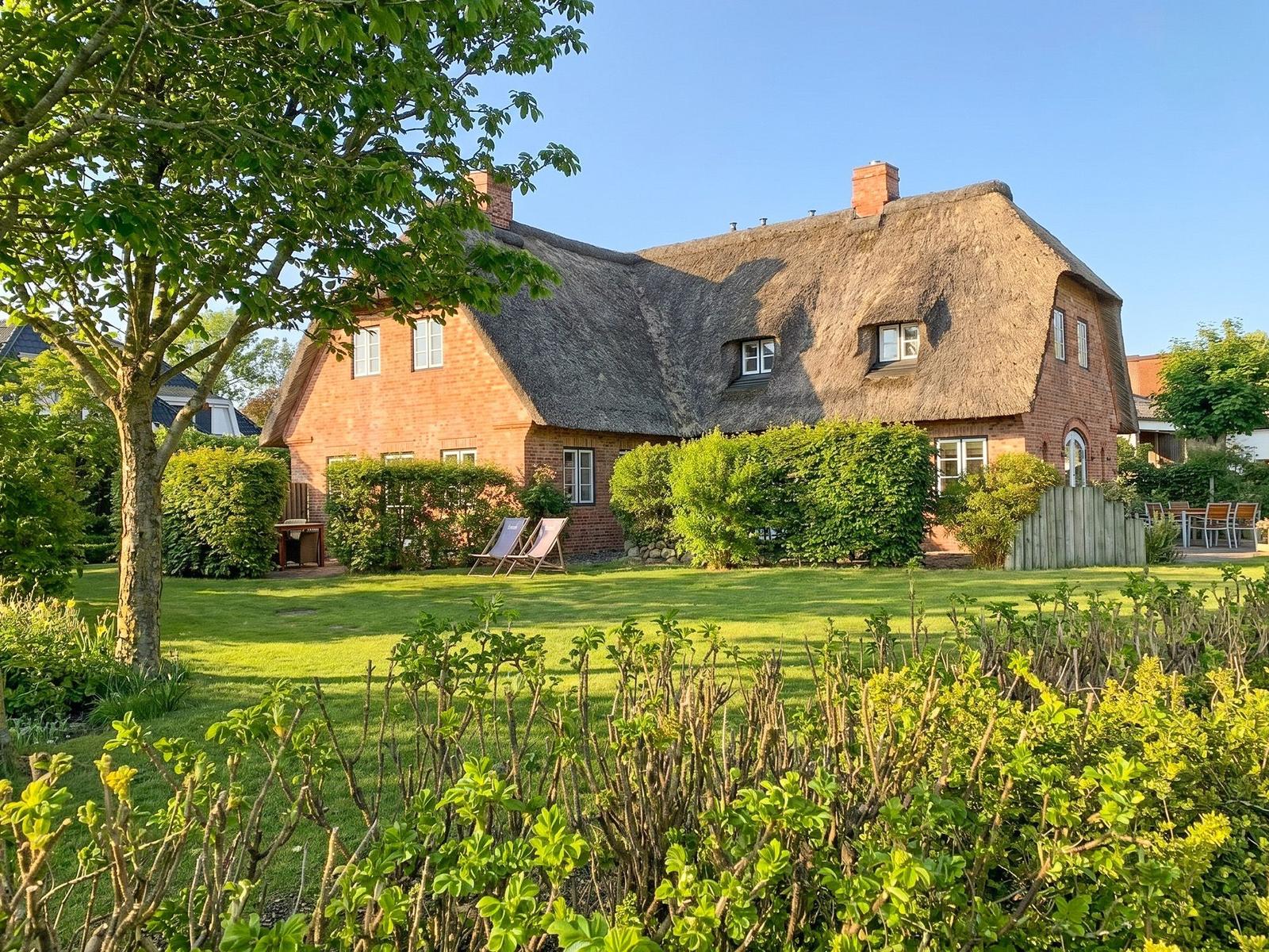 Red brick house with thatched roof, surrounded by greenery and trees.