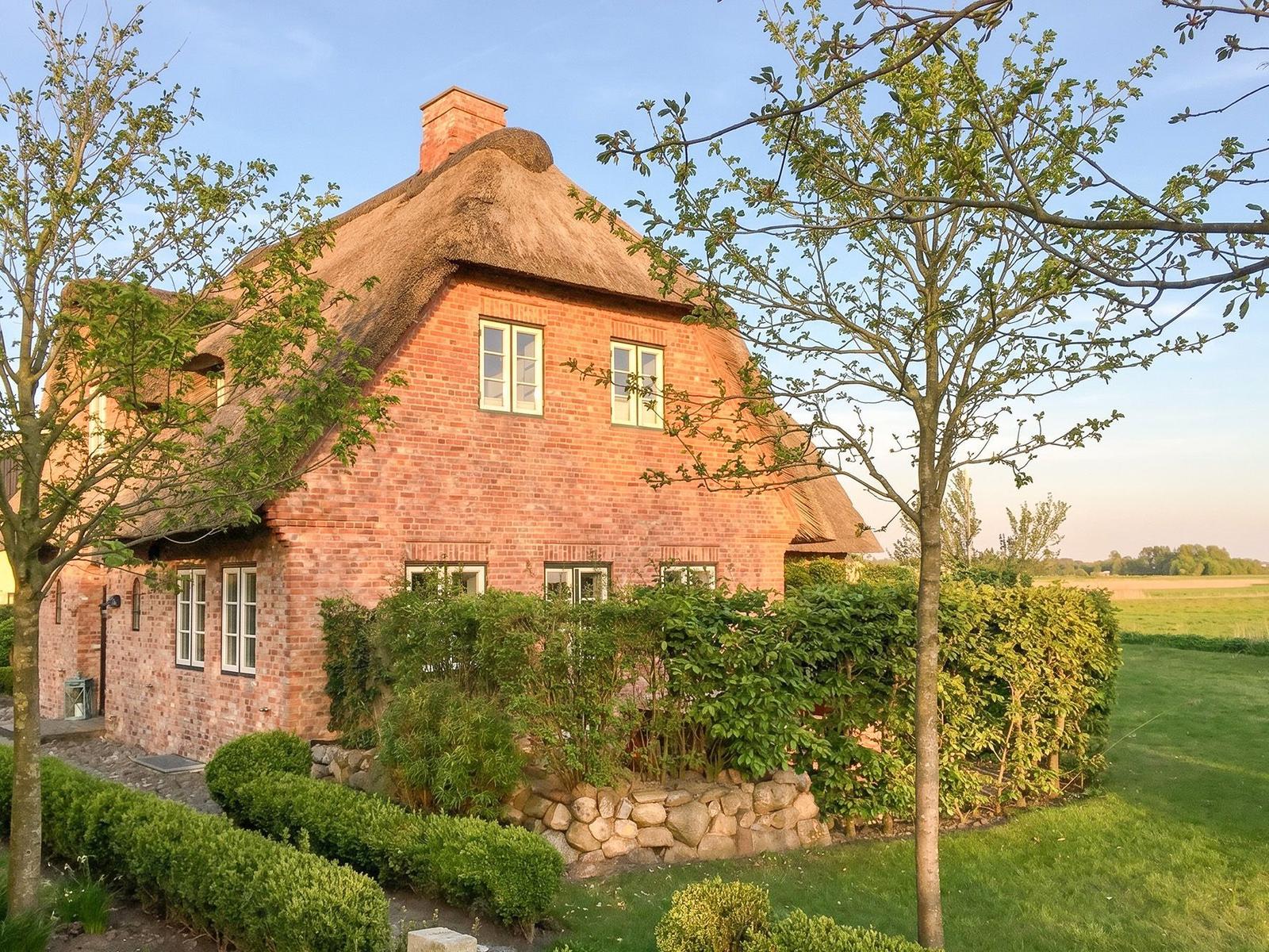 Red brick house with thatched roof, surrounded by greenery and trees.