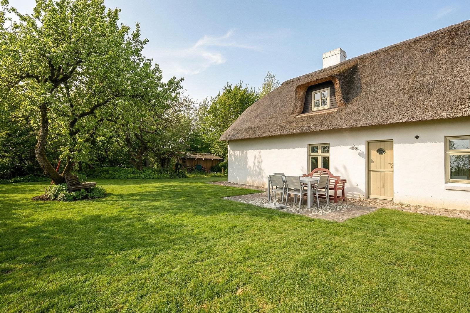 Haus mit Strohdach, Terrasse und Garten. Holztisch mit Stühlen. Bäume und Grasfläche.
