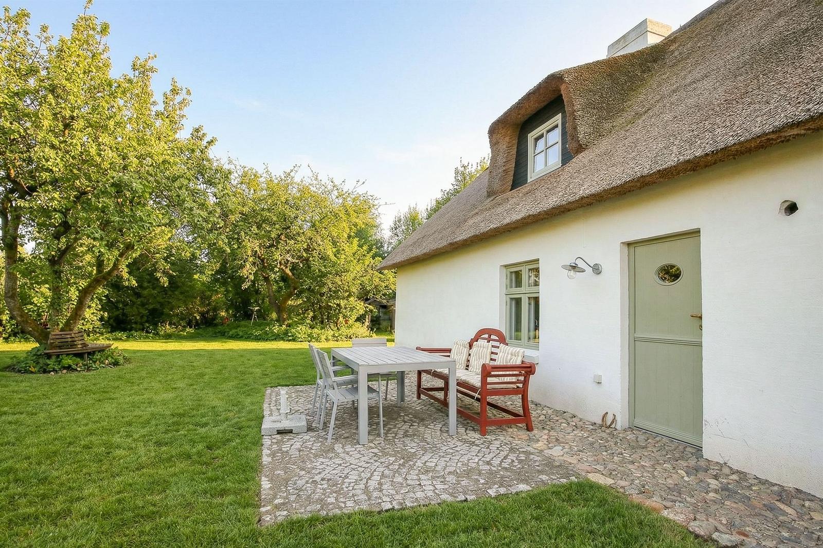 Haus mit Strohdach, Terrasse mit Tisch, Stühlen und Bank. Grünfläche und Bäume im Hintergrund.