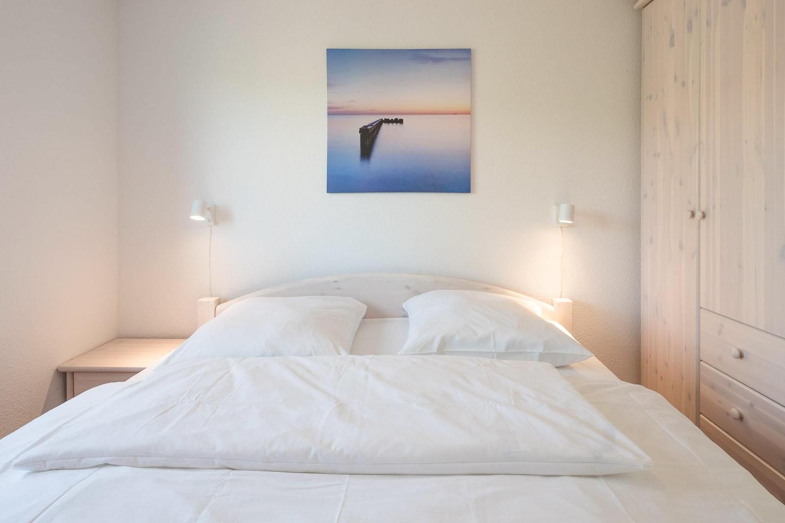 Schlafzimmer mit Bett, Nachttisch und Kleiderschrank. Wandbild zeigt einen Steg am Wasser.