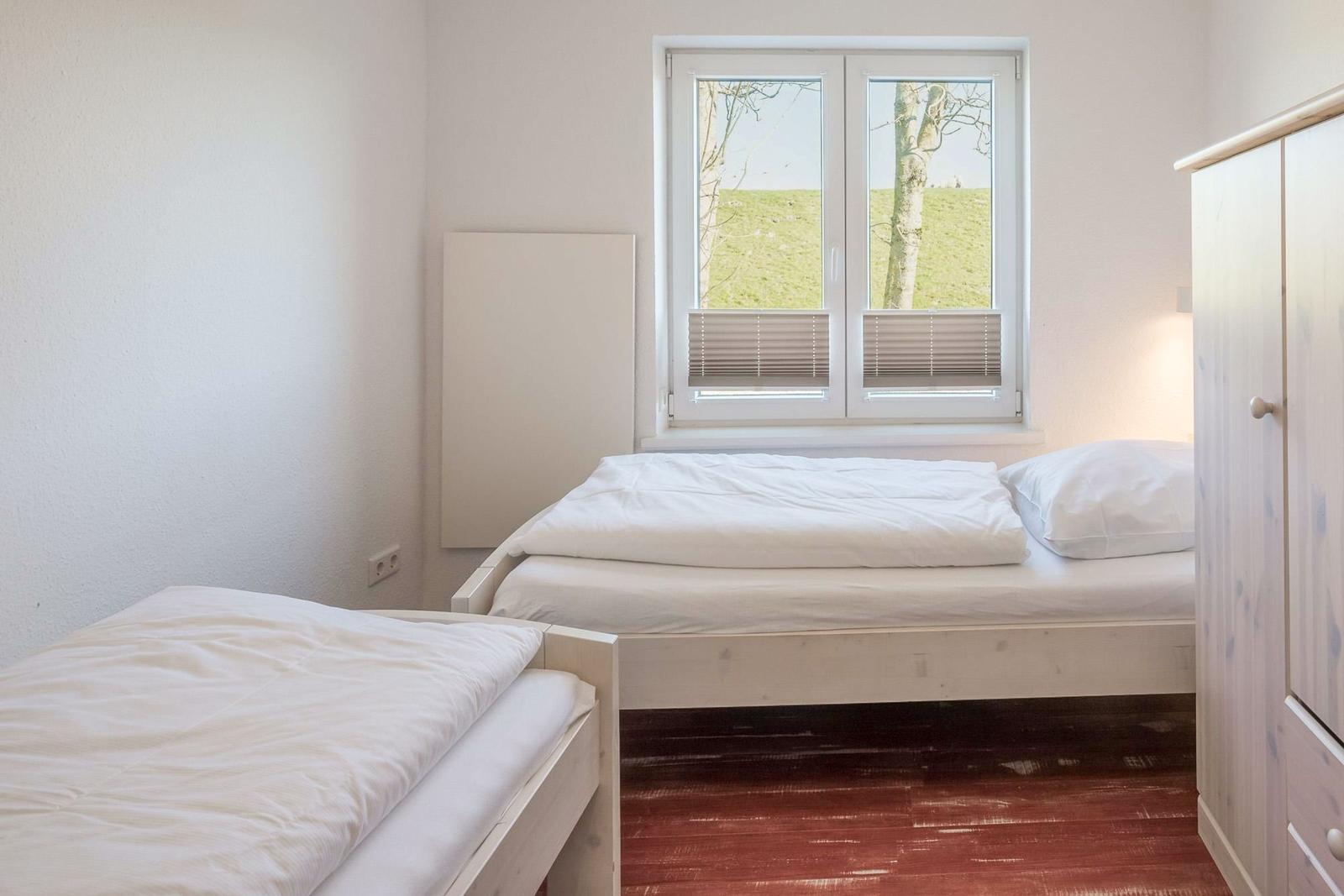 Zwei Betten mit weißer Bettwäsche, Fenster mit Blick auf Gras und Baum, Schrank und Holzboden.