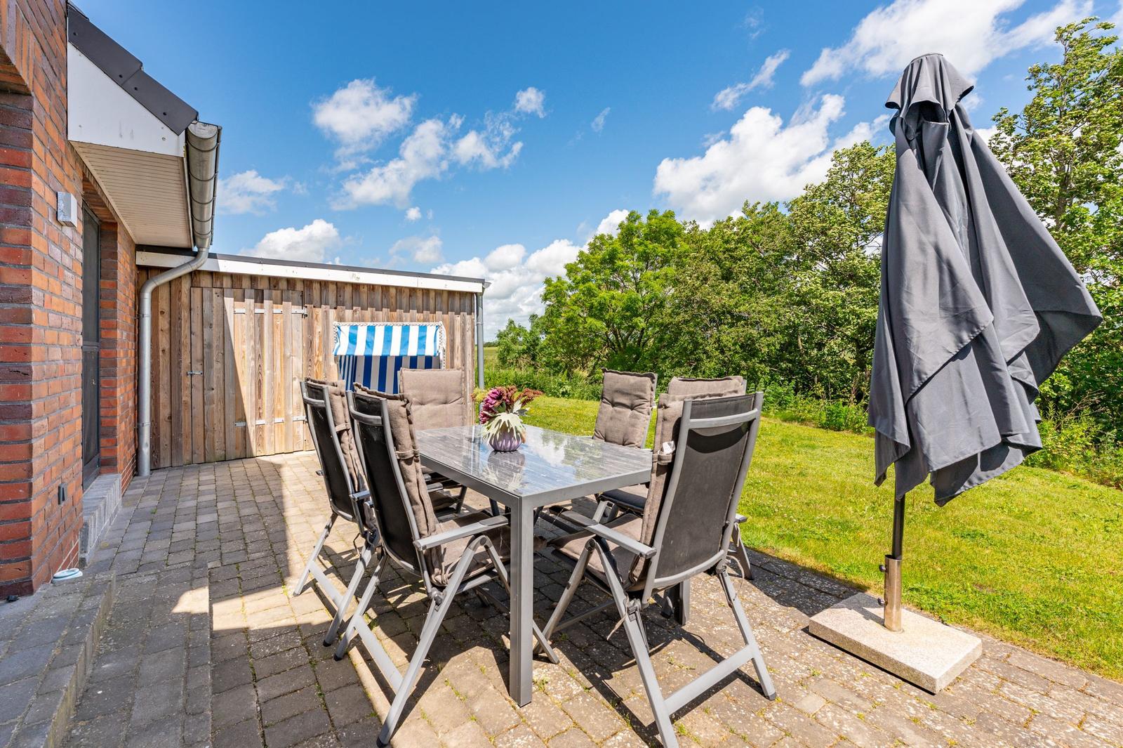 Terrasse mit Tisch, Stühlen und geschlossenem Sonnenschirm. Hintergrund: Holzhaus und Grünflächen.