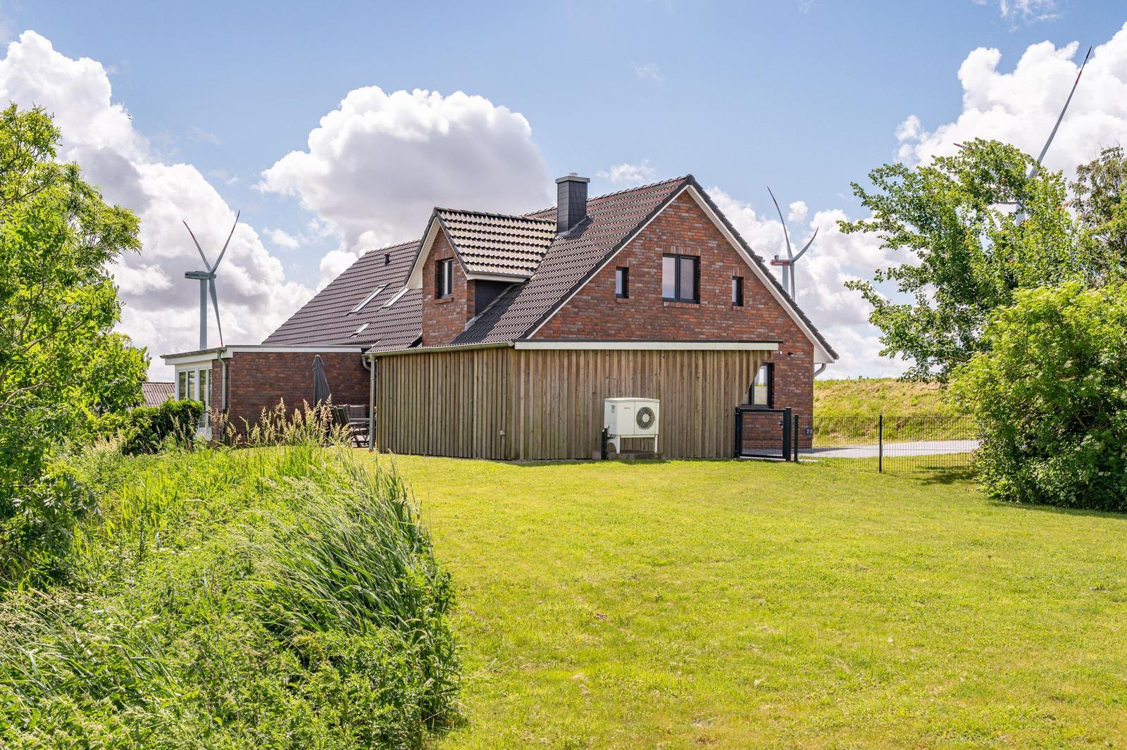 Haus mit Holzverkleidung und Dachgauben. Grünfläche und Windräder im Hintergrund.