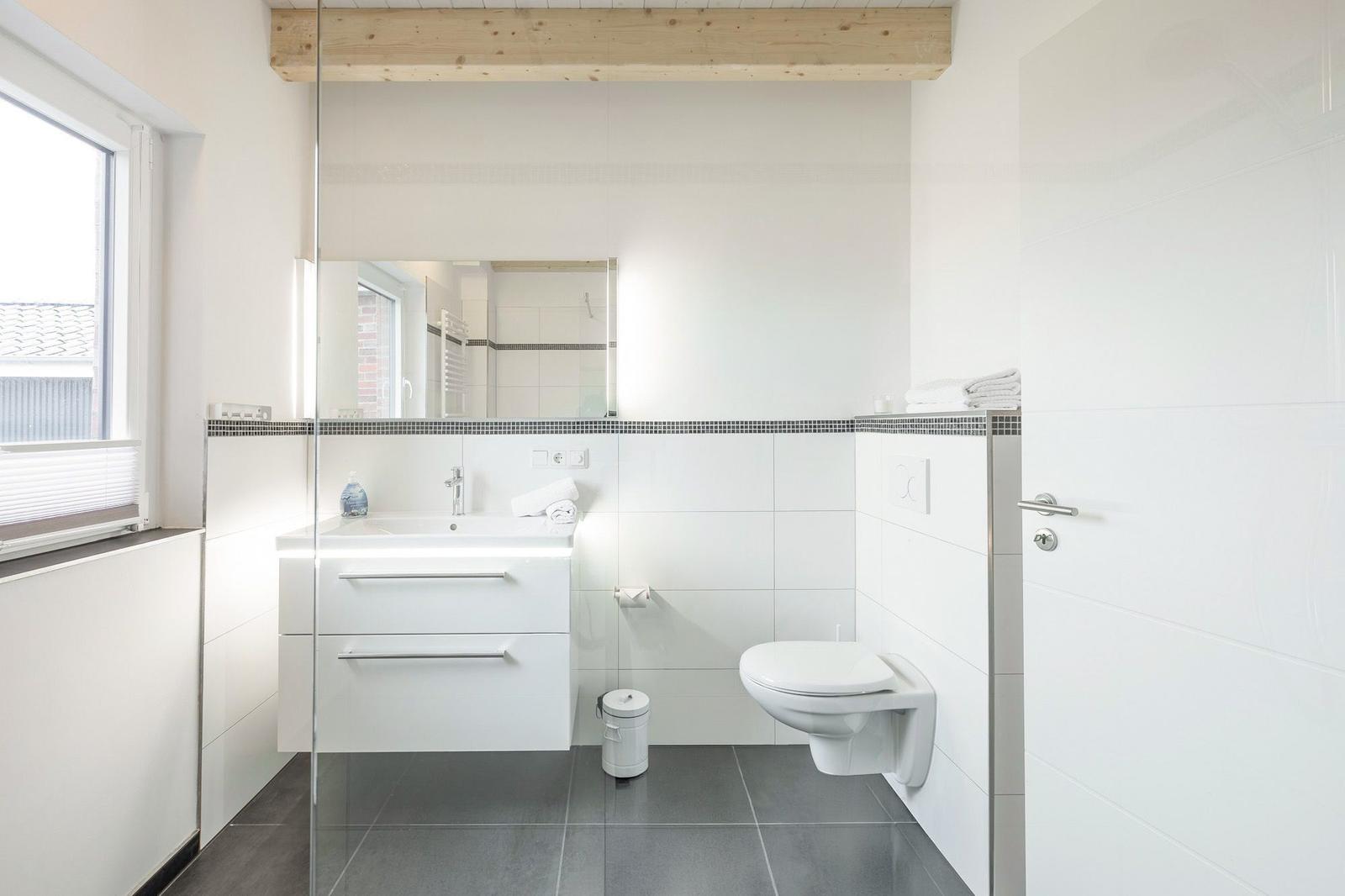 Badezimmer mit Waschbecken, Toilette und Glasdusche. Weiße Fliesen und Holzbalken.