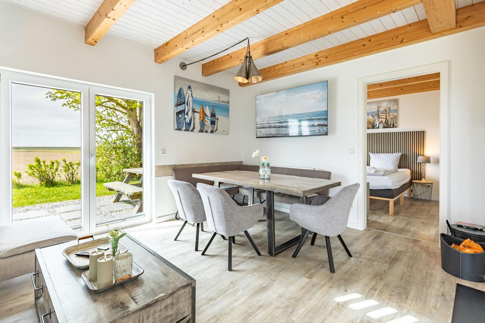 Esszimmer mit Tisch, Stühlen und Blick auf Garten. Schlafzimmer mit Bett und Holzdecke sichtbar.
