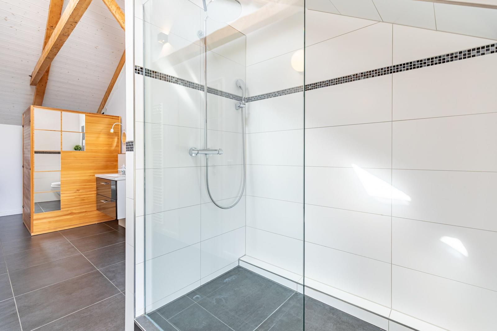 Dusche mit Glaswand, weiße Fliesen und Holzdeckenkonstruktion. Toilettenbereich mit Holzumrandung.