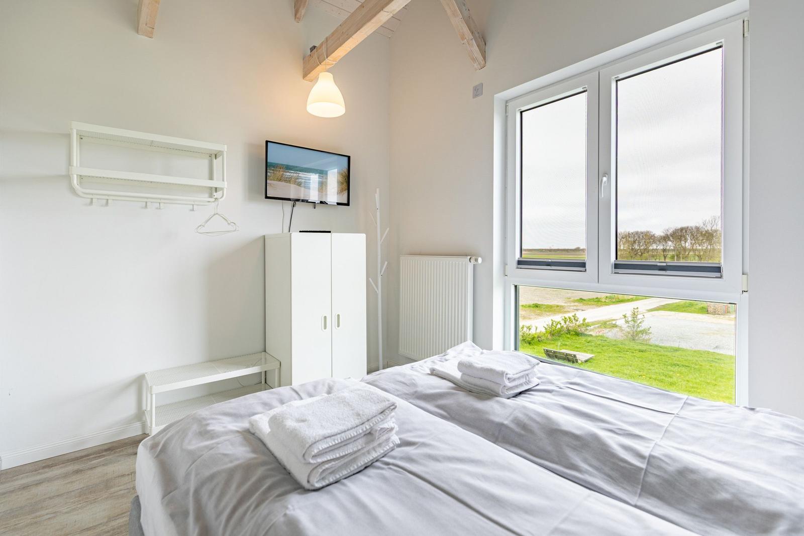 Schlafzimmer mit Bett, Kleiderständer und TV. Große Fenster mit Blick auf Grünfläche.