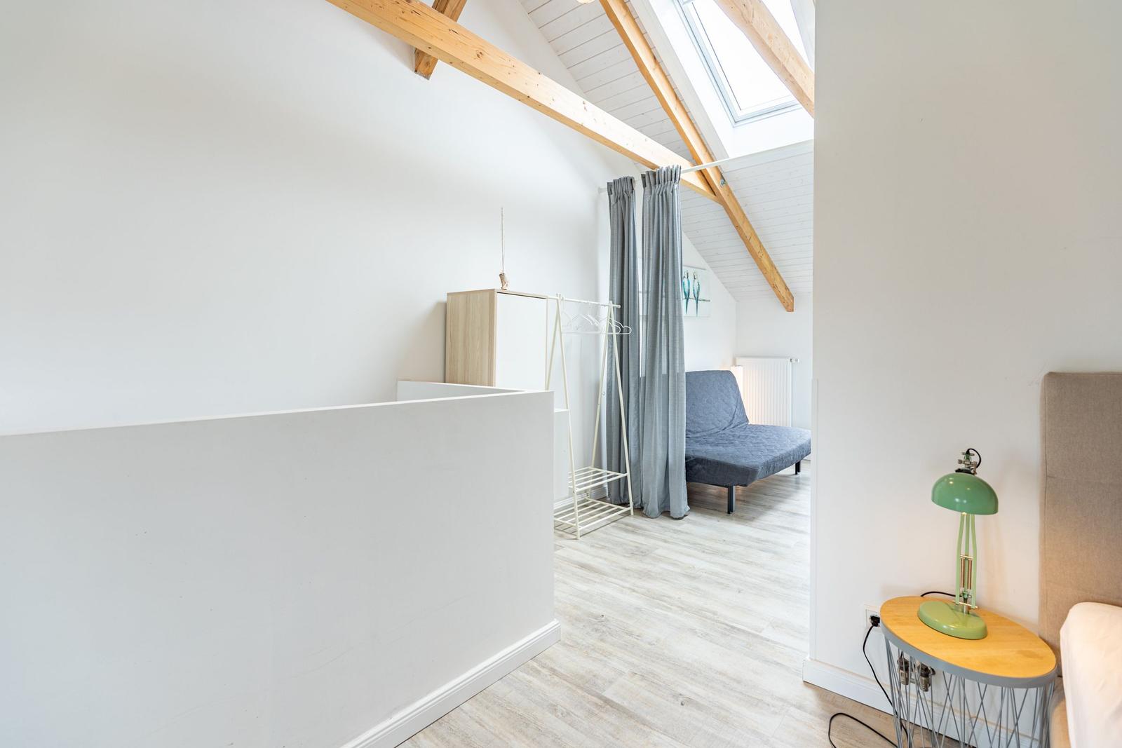 Schlafzimmer mit Dachschräge, Holzbalken und Fenster. Neben einem Bett steht ein Nachttisch mit Lampe.