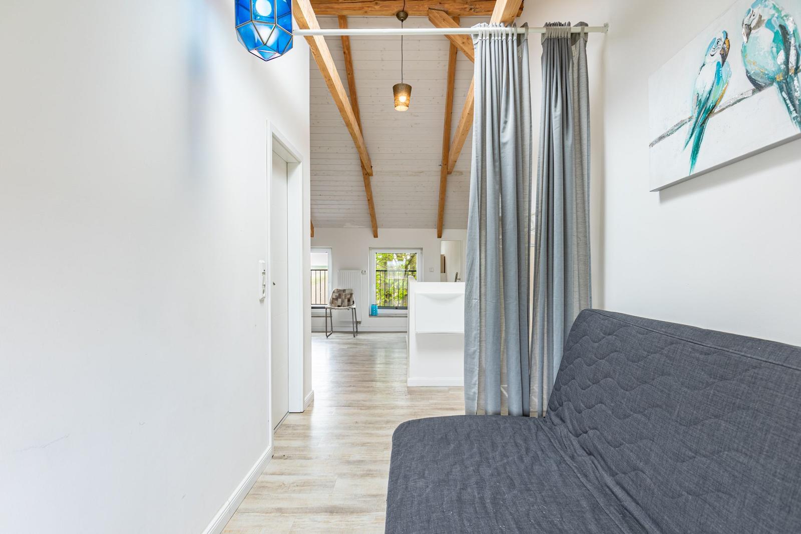 Ein Raum mit grauem Sofa, grauen Vorhängen und einer blauen Lampe. Holzbalken und ein Wandbild sind sichtbar.