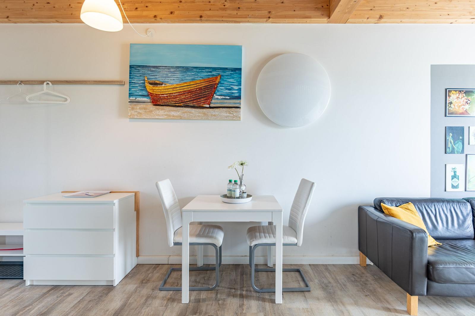 Ein Essbereich mit Tisch, zwei Stühlen und einem Sofa. Wandbild zeigt ein Boot.