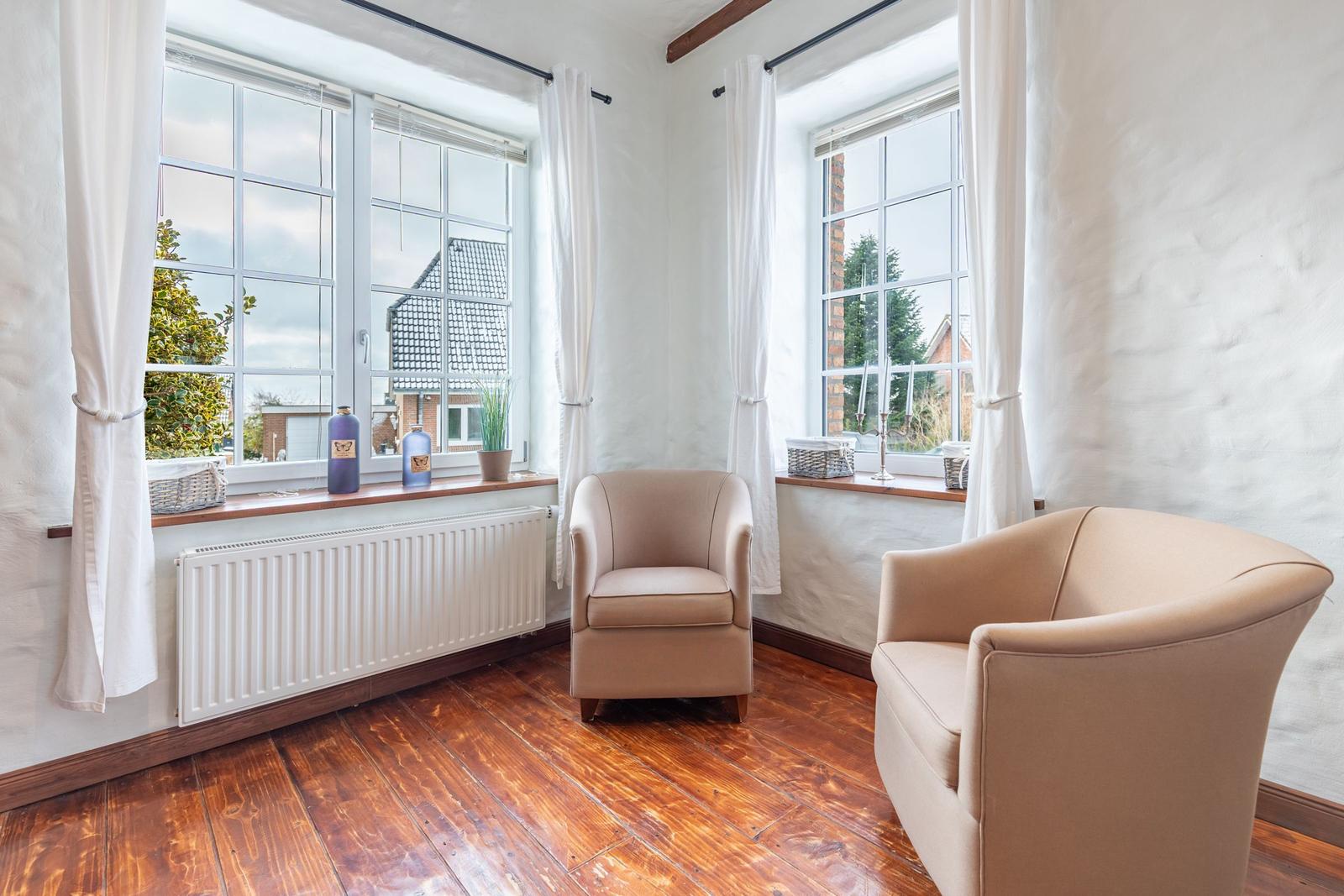Two beige armchairs are in a room with hardwood floors and large windows.