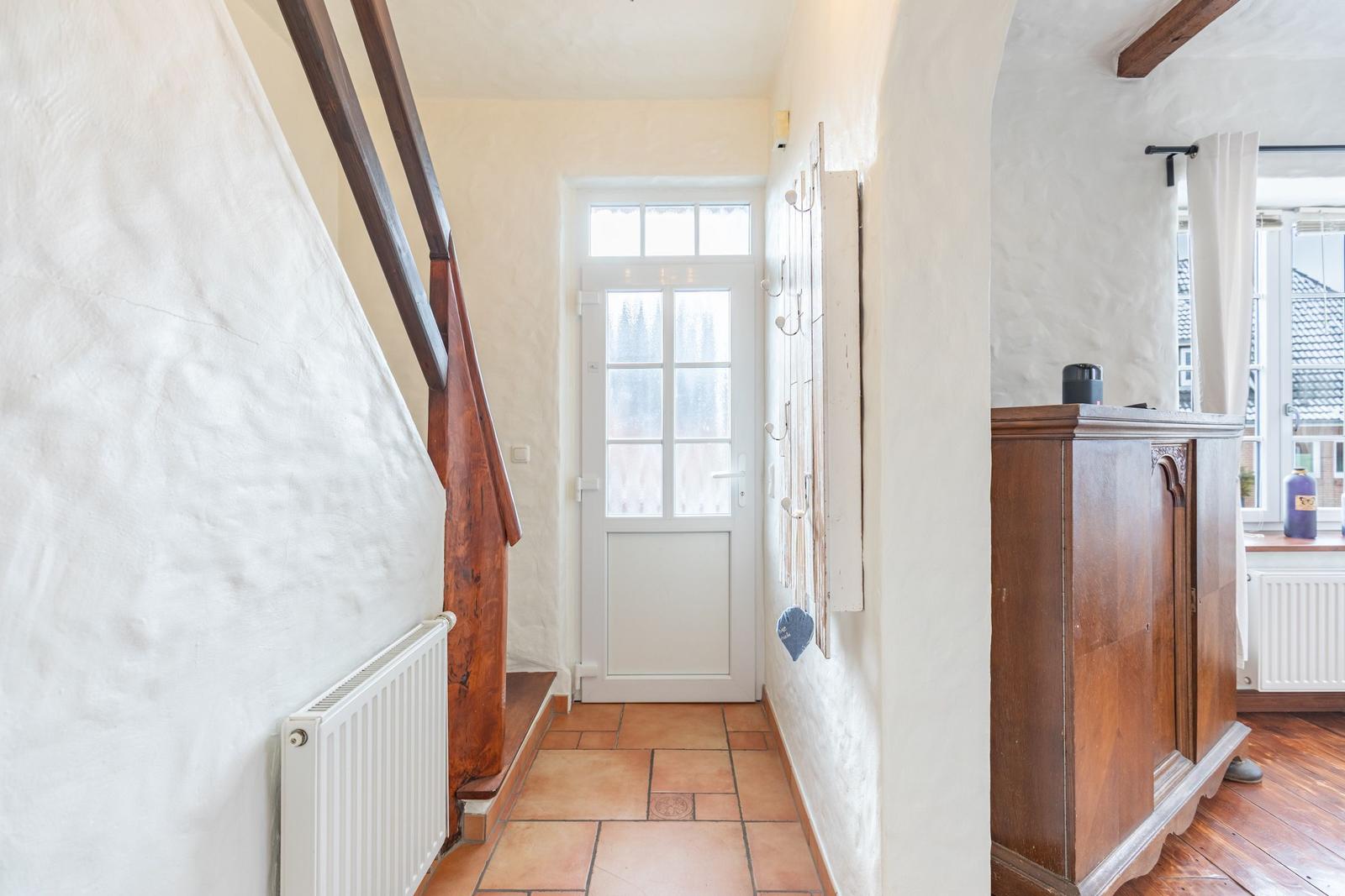 Entrance with wooden staircase, white door, and wooden cabinet. Terracotta floor and white walls.