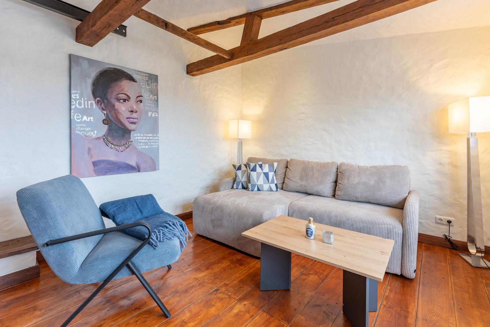 Living room with sofa, armchair, table, and wall art under wooden beams.