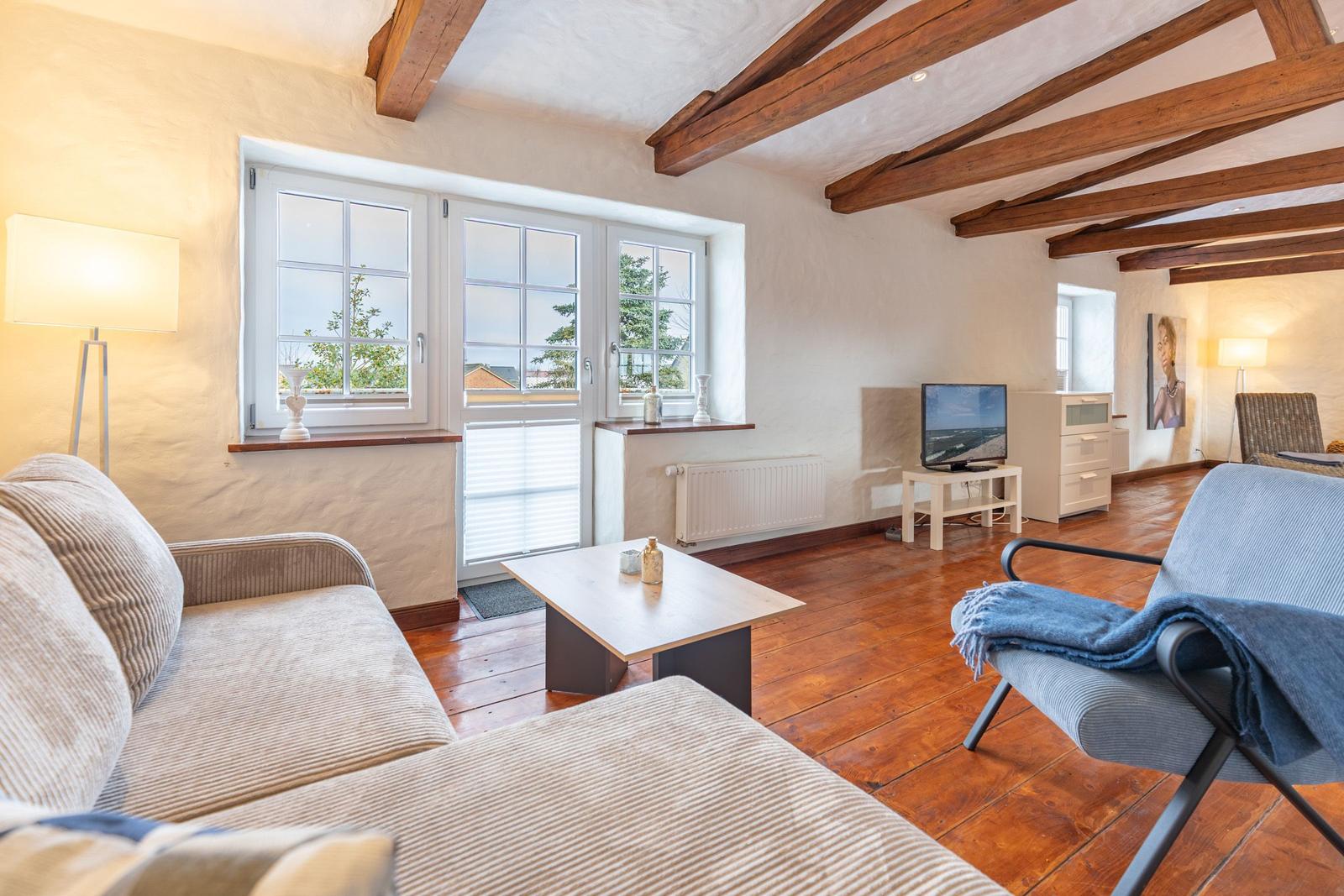 Living room with wooden beams, sofa, armchair, and TV. Wooden floor and windows with view.