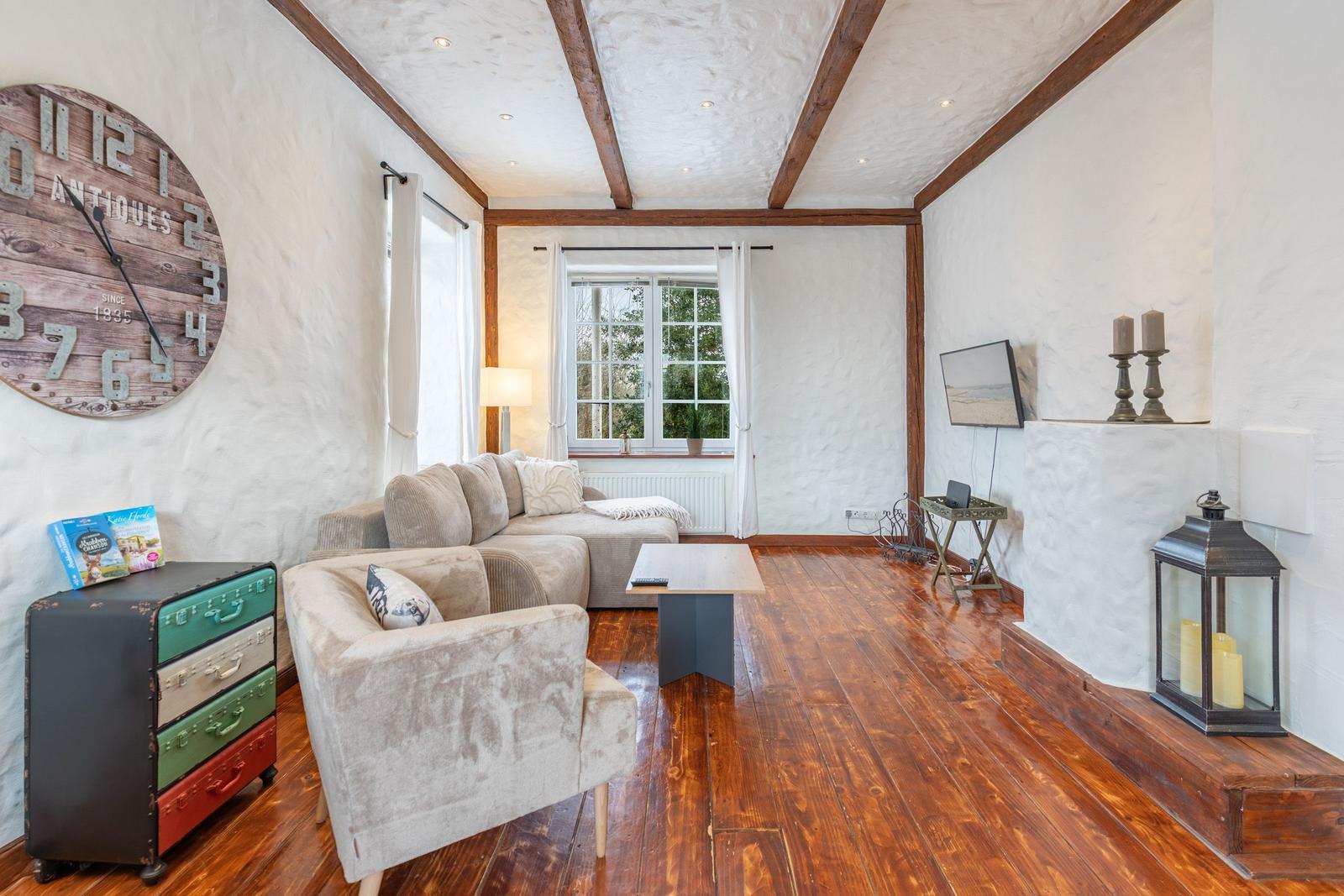 Living room with wooden floor, sofa, armchair, and wall clock. Window with garden view.