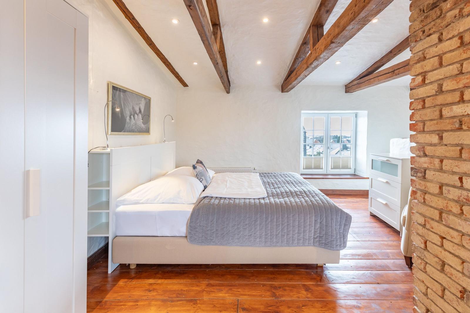 Bedroom with wooden floor, exposed beams, window, and white bed.