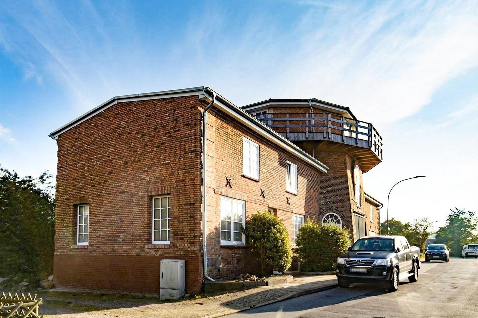 Red brick house with balcony and windows, parked cars in front of the facade.
