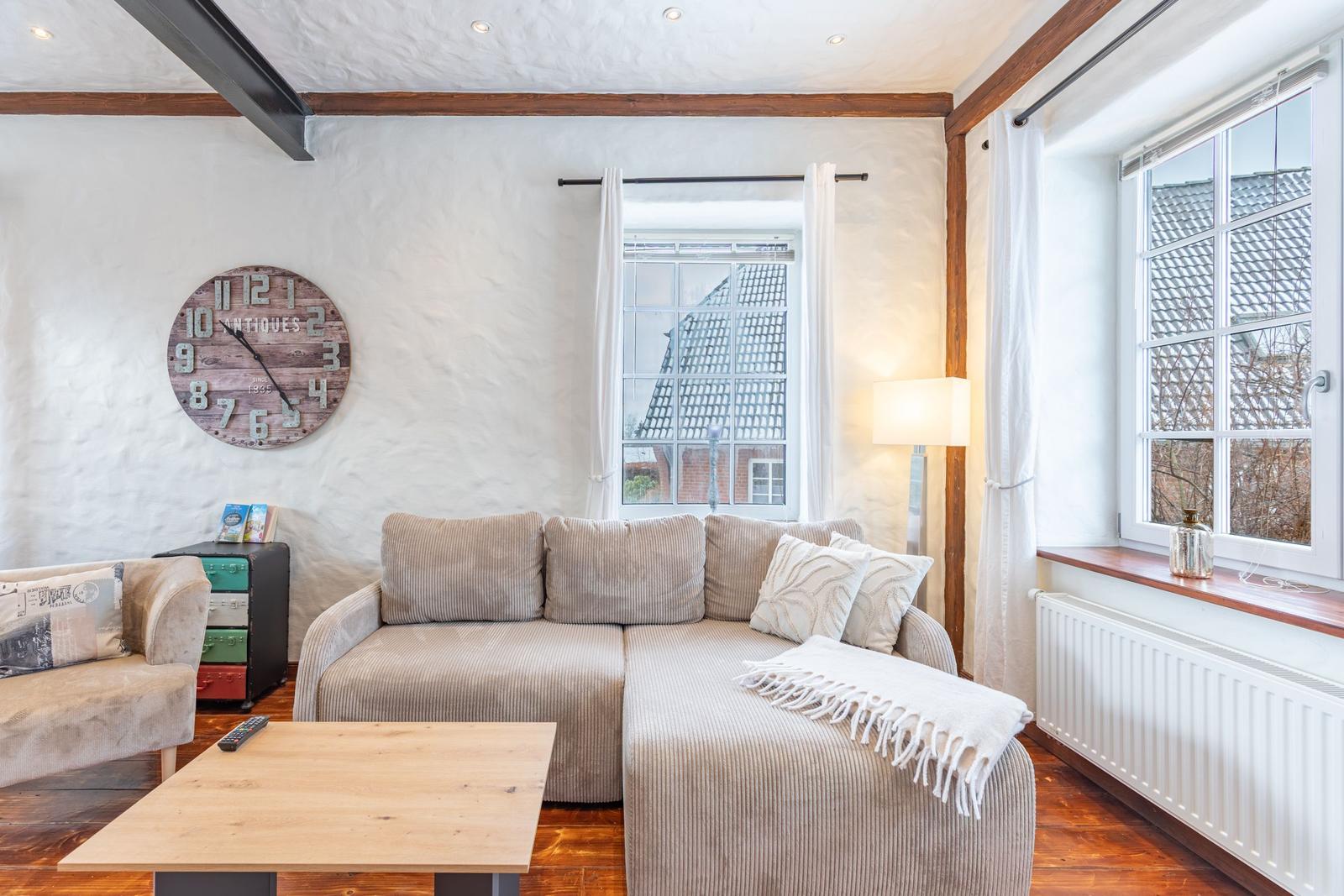 Living room with sofa, table, and wall clock. Wooden windows and beams.