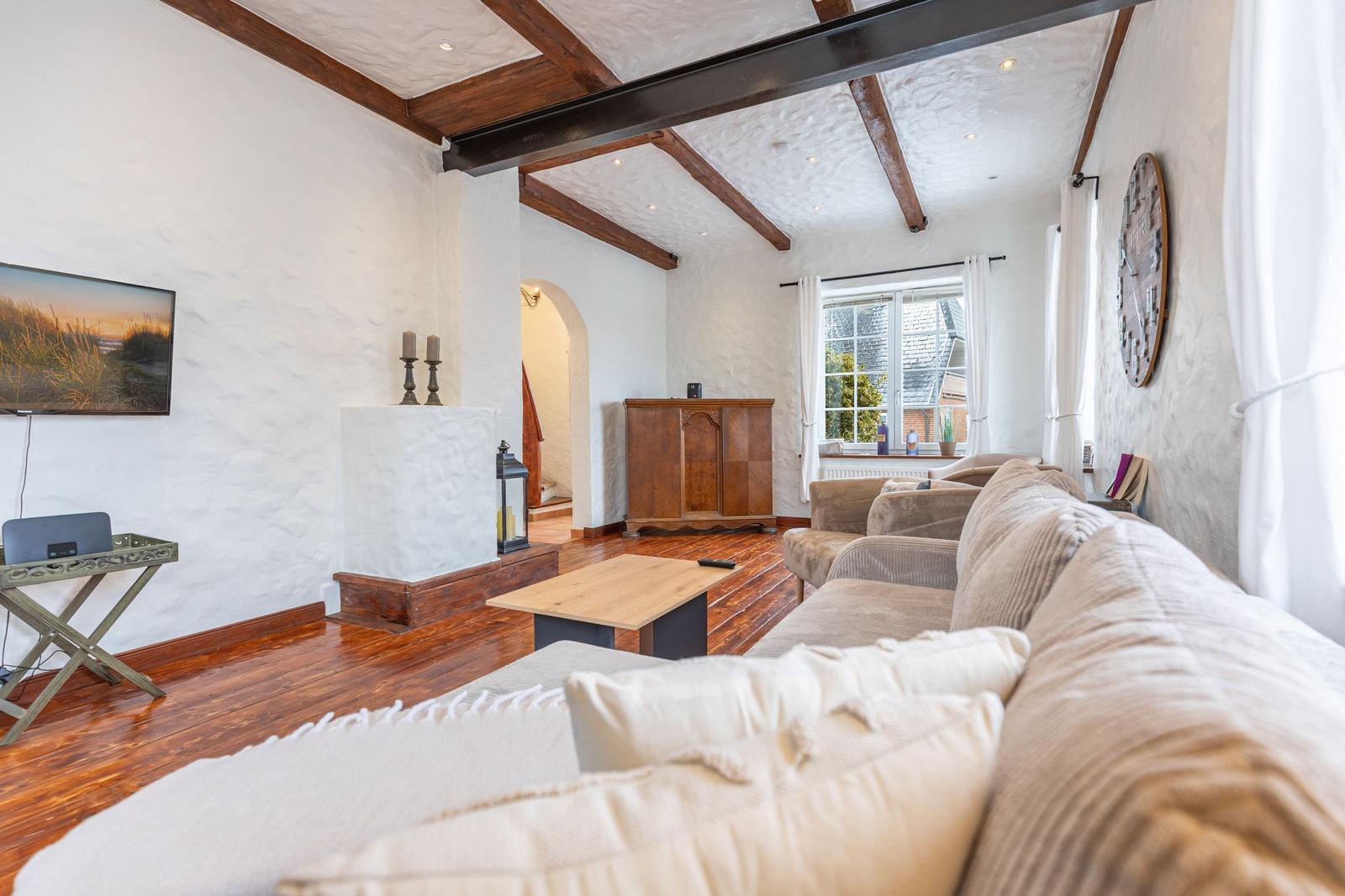 Living room with wooden floor, sofa, TV, and fireplace. Beams and window visible.