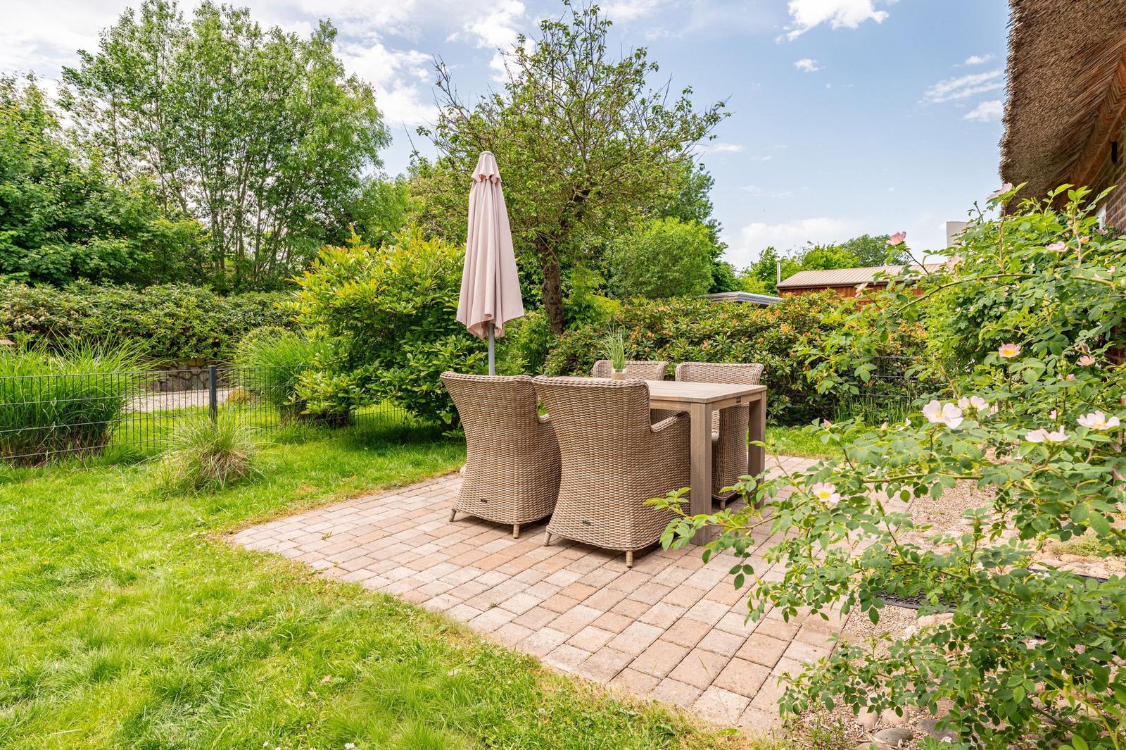 Terrace with table, chairs, and closed umbrella. Greenery and garden house in background.