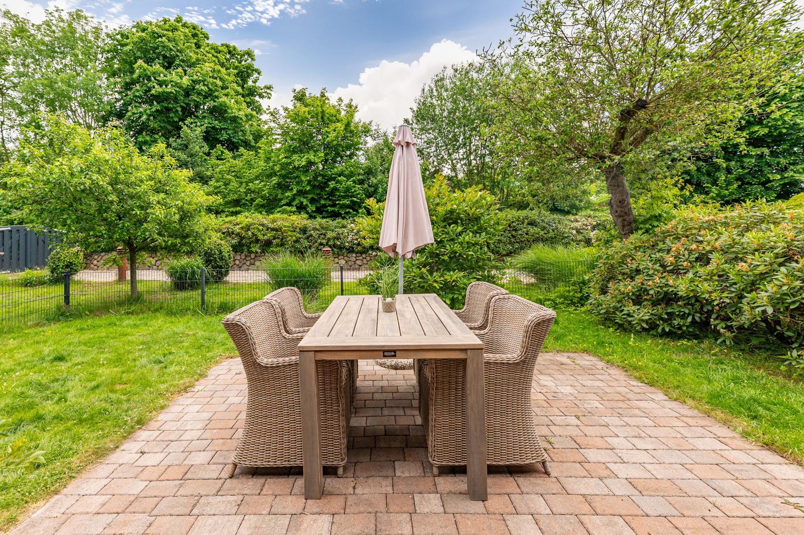 Garden with table, four chairs, and closed umbrella on stone paving.
