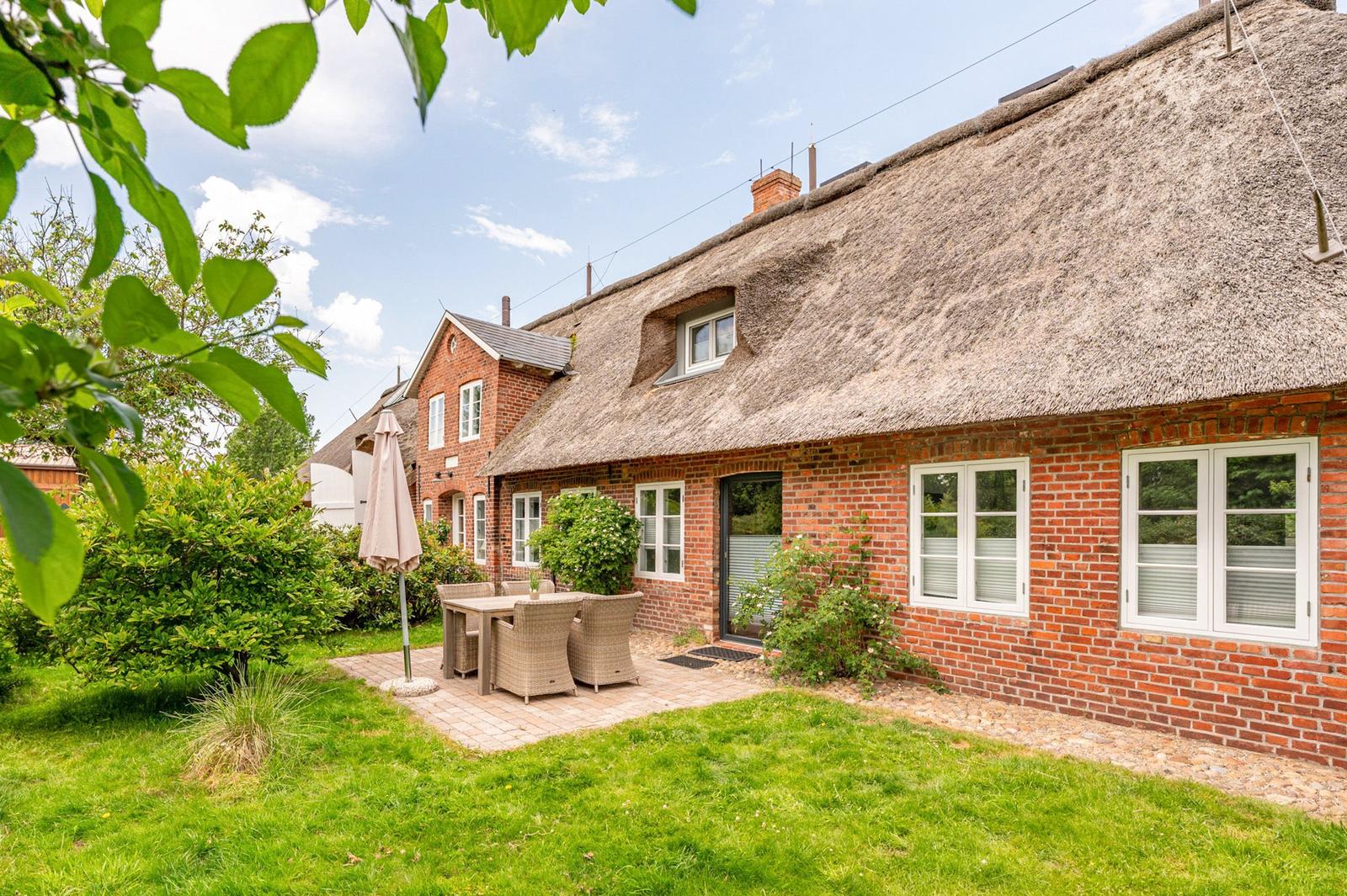 House with thatched roof, garden with table and chairs, green grass and trees.