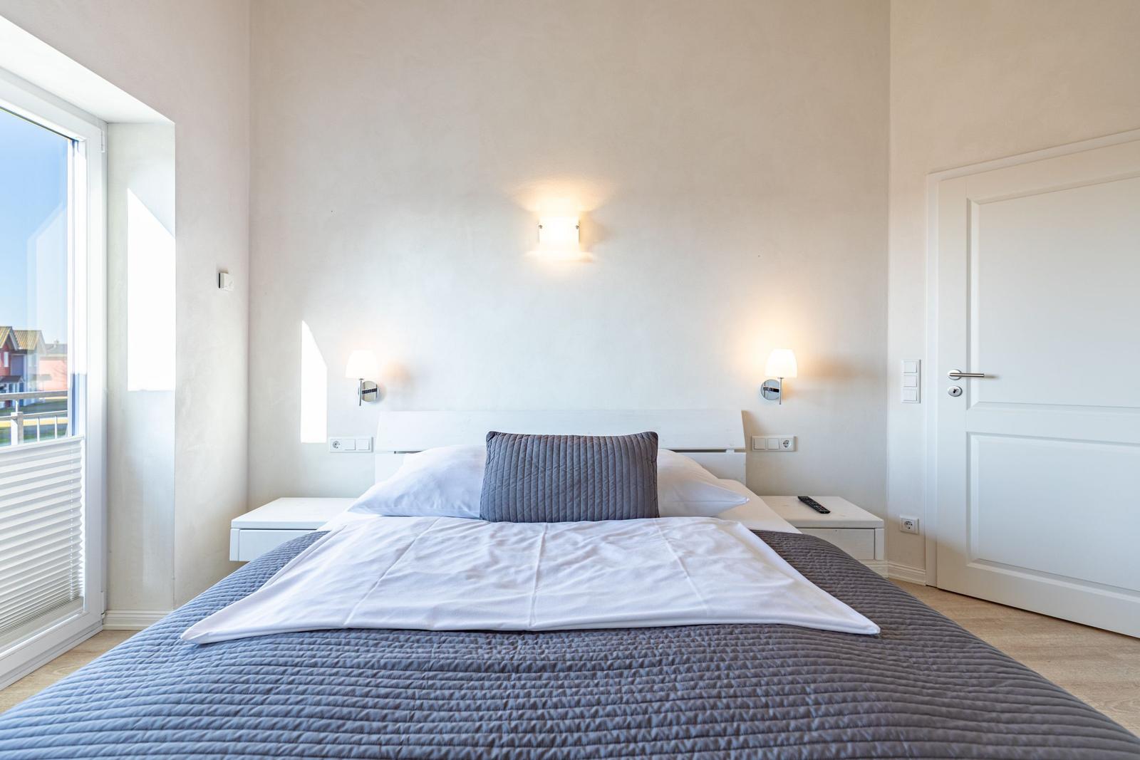 Bedroom with bed, nightstands, and window. Wall lamps and door are visible.