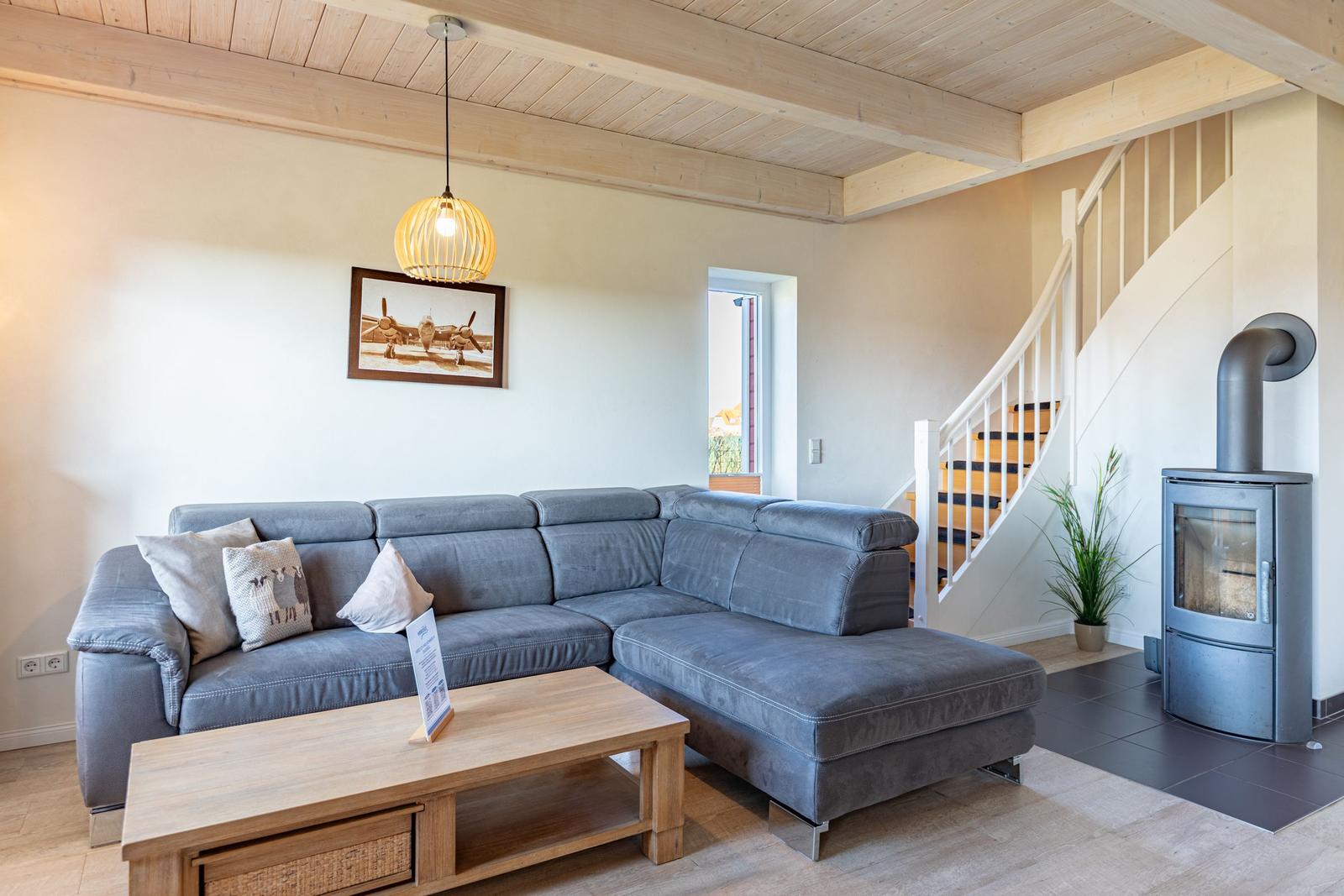 Living room with gray sofa, wooden table, and wood stove. Light walls and wooden ceiling.