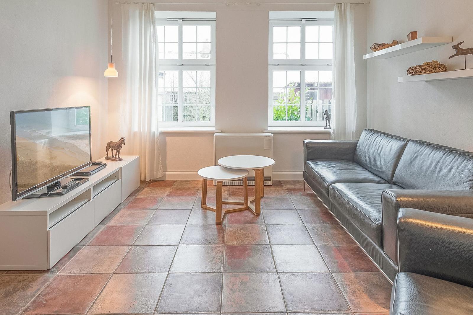 Ein Wohnzimmer mit Sofa, Fernseher und zwei Fenstern. Der Boden ist mit Fliesen ausgelegt.