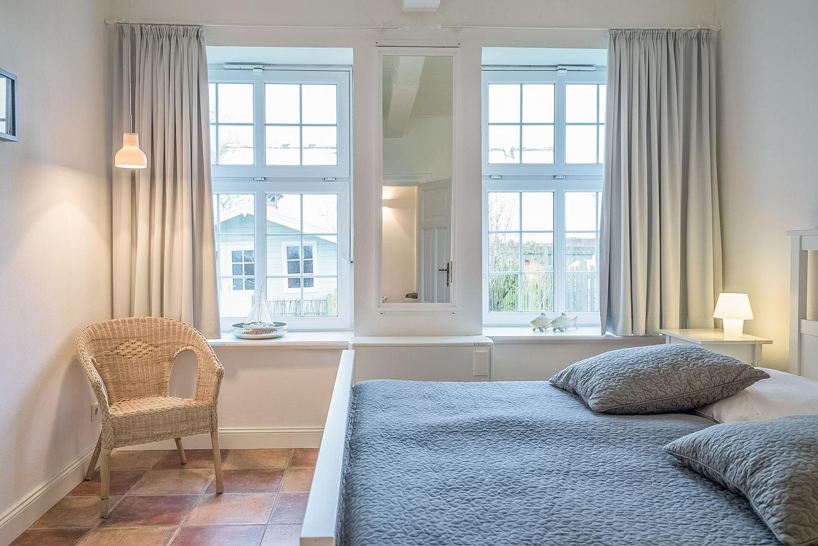 Schlafzimmer mit Bett, Fenstern und Stuhl. Fenster zeigen Außenansicht.