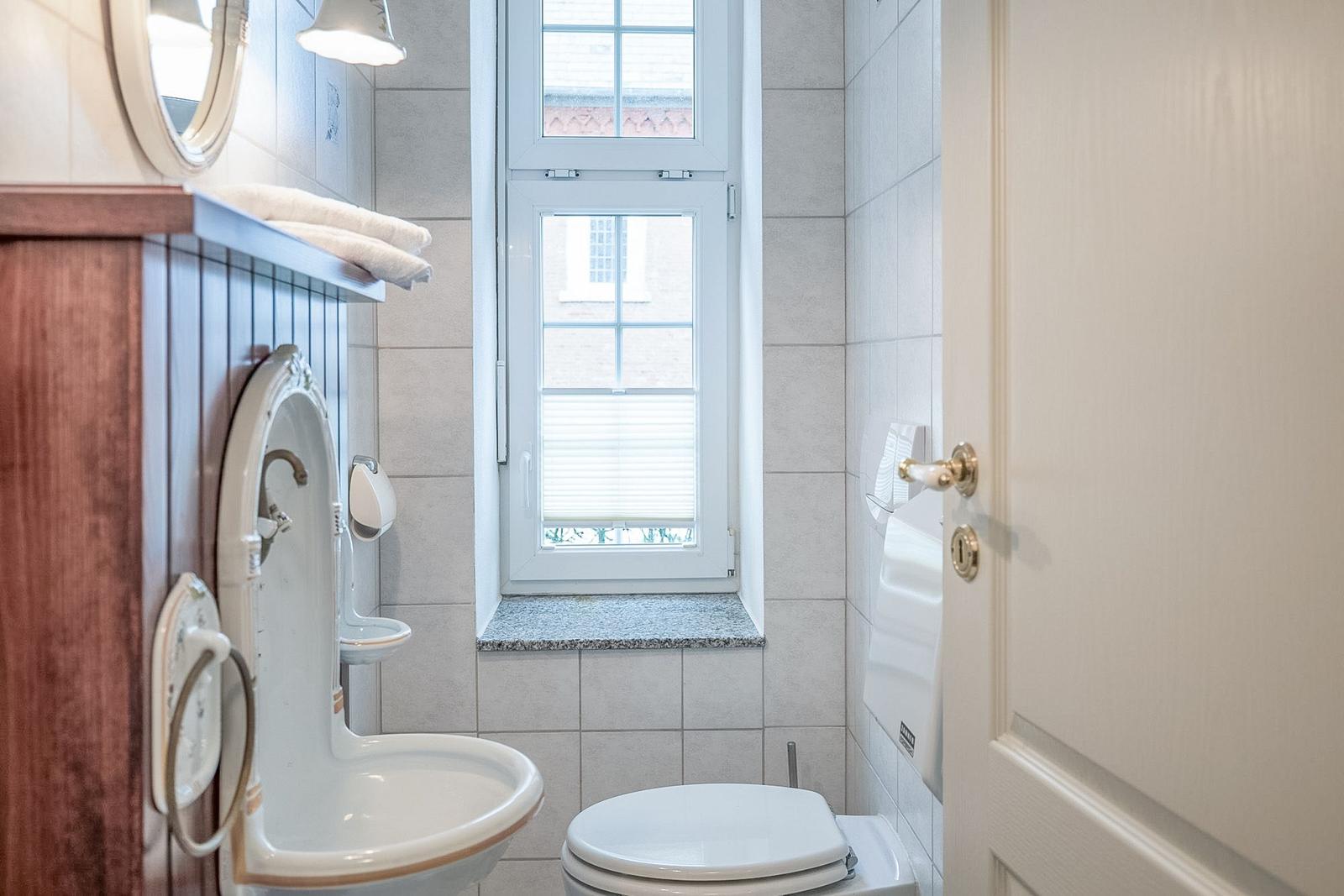 Badezimmer mit Toilette, Waschbecken und Fenster. Holzregal und Spiegel sichtbar.
