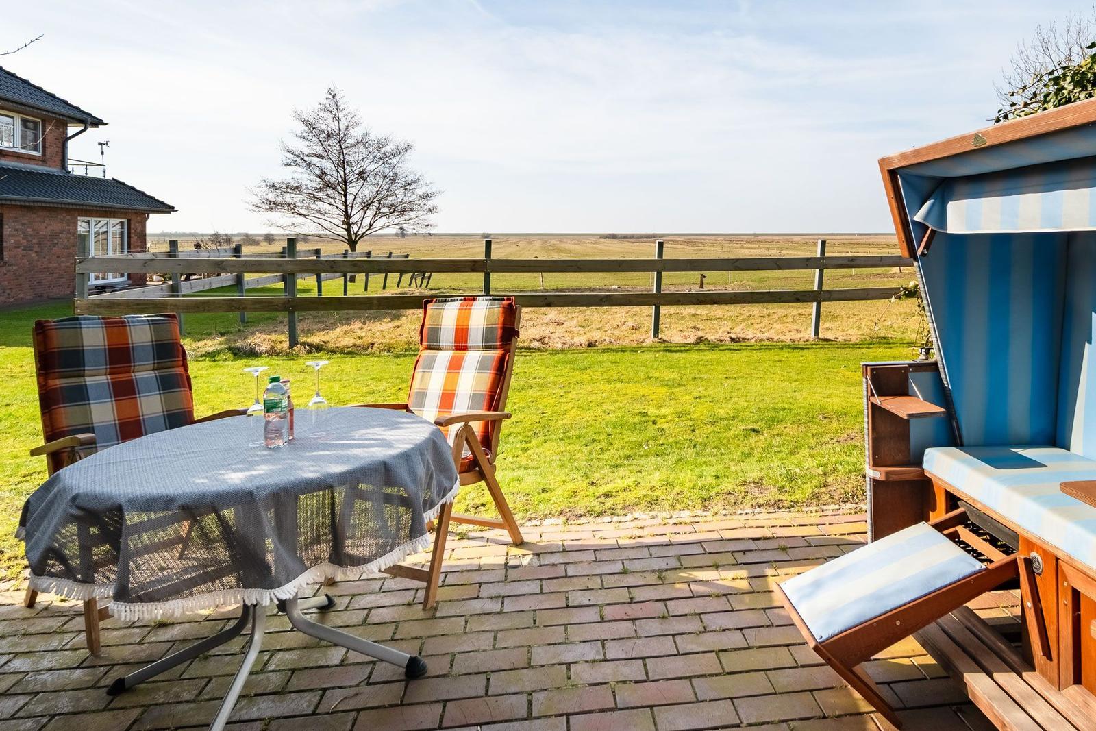 Terrasse mit Tisch, zwei Stühlen und Strandkorb. Blick auf grünes Feld und Holzzaun.