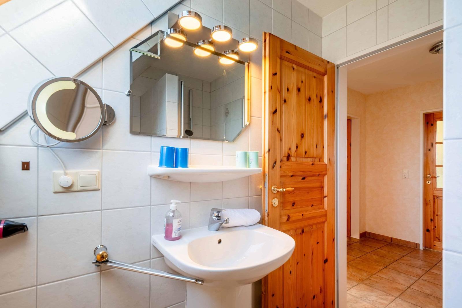 Bad mit Waschbecken, Spiegel und Holztür. Dusche sichtbar im Hintergrund.
