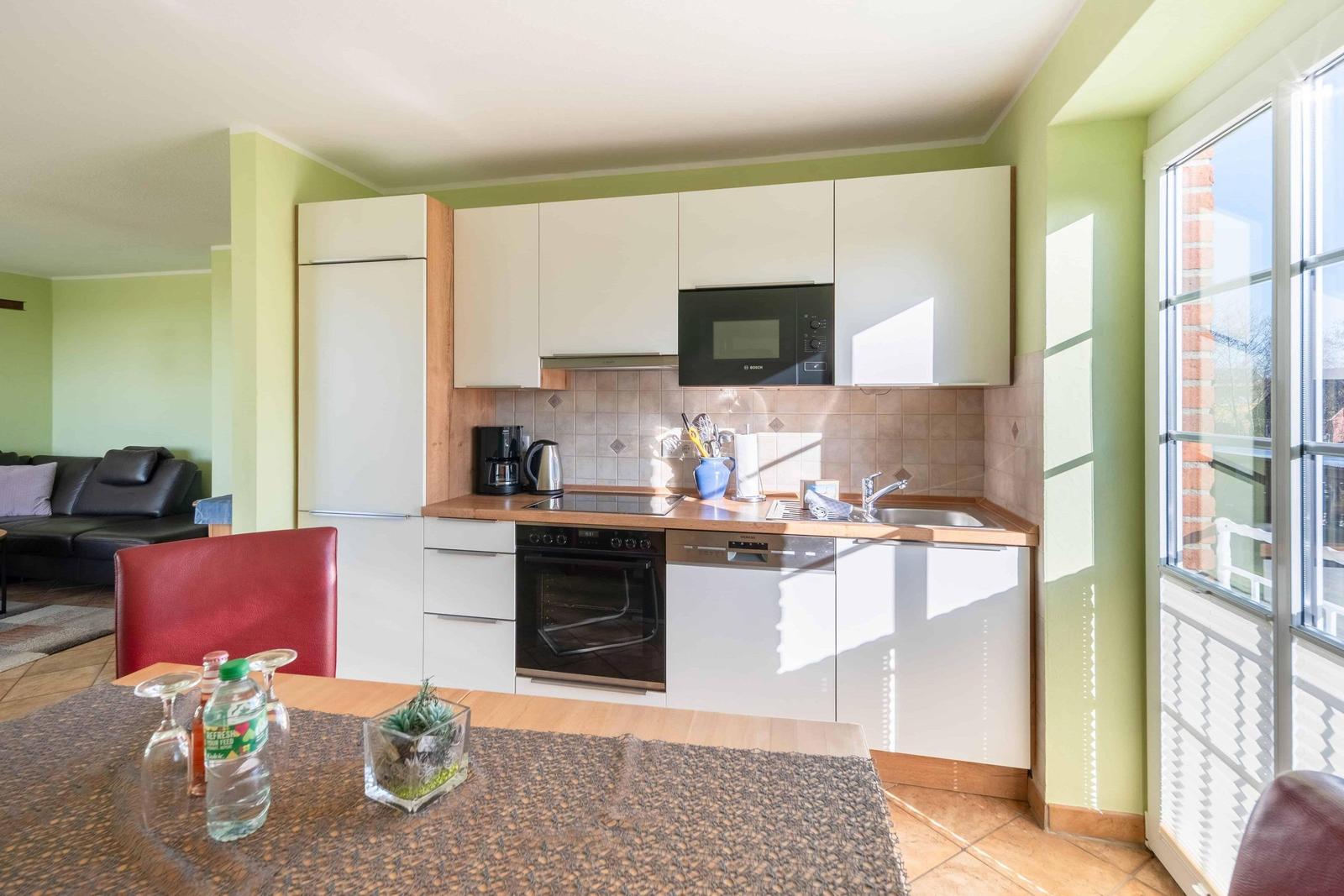 Küche mit weißen Schränken, Herd, Spüle und Fenster. Esstisch mit roter Stuhl.
